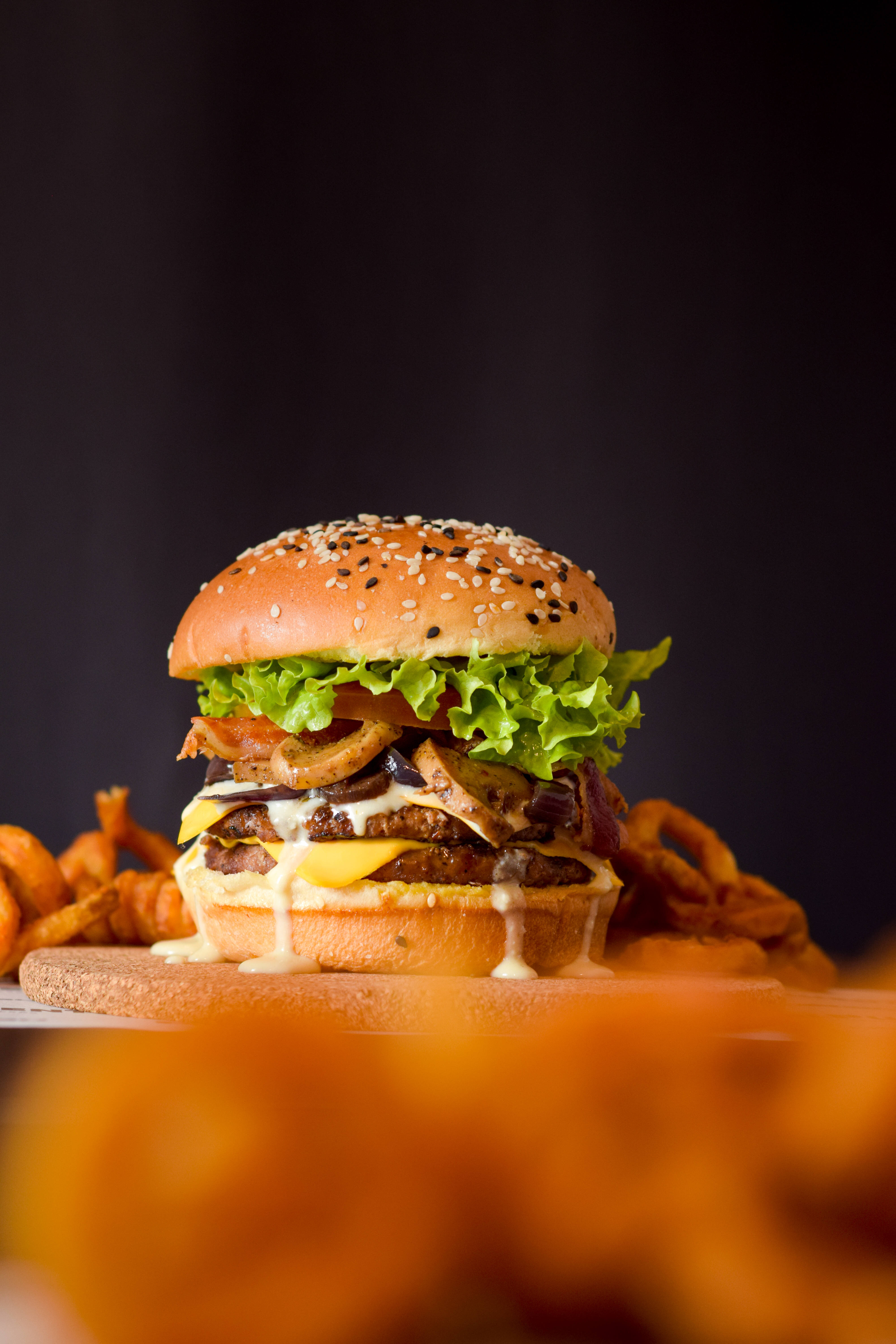Food Photography 桌布 壁紙 — 一個多汁的漢堡，配上融化起司、培根和生菜，放在木板上，周圍是酥脆的洋蔥圈，背景深色。