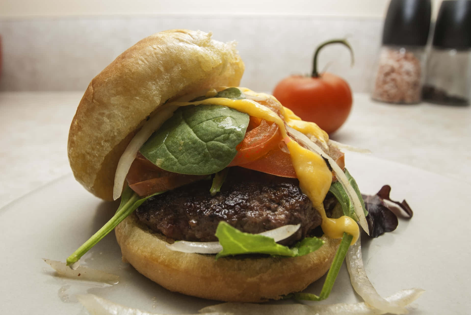 hamburger 桌布 壁紙 — 一個多汁的漢堡，配有融化起司、番茄、菠菜和洋蔥，放在白色盤子上，背景有一個新鮮番茄。