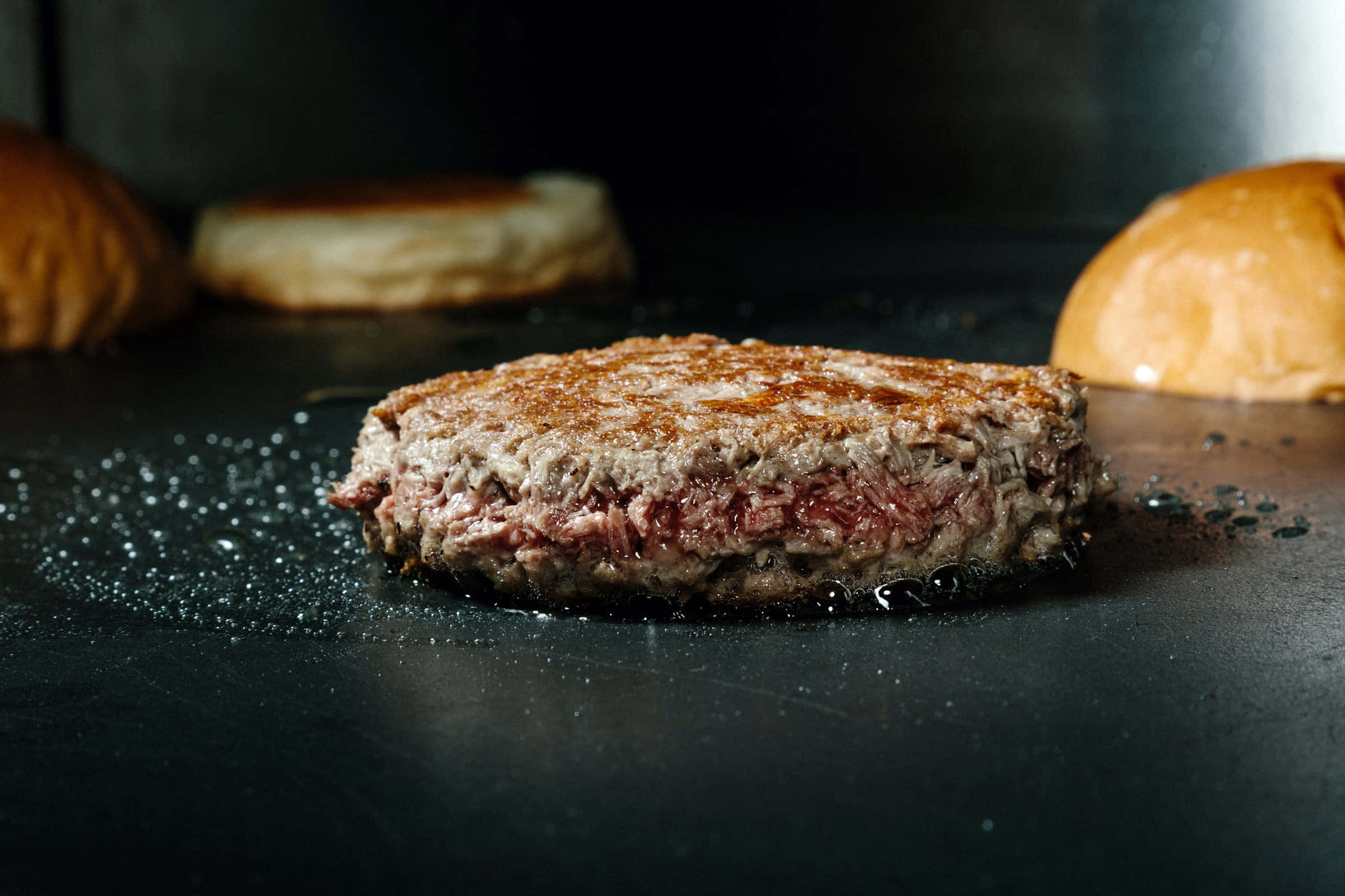 fast food 桌布 壁紙 — 一個多汁的漢堡肉餅在黑色烤盤上滋滋作響，旁邊有漢堡麵包，特寫鏡頭。