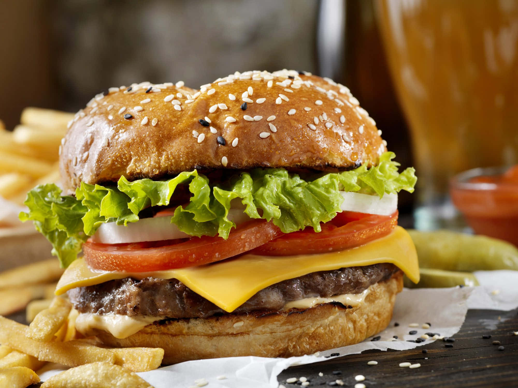 hamburger 桌布 壁紙 — 一個多汁的芝麻漢堡，配上生菜、番茄、起司和洋蔥，旁邊是酥脆的薯條，放在質樸的木桌上。