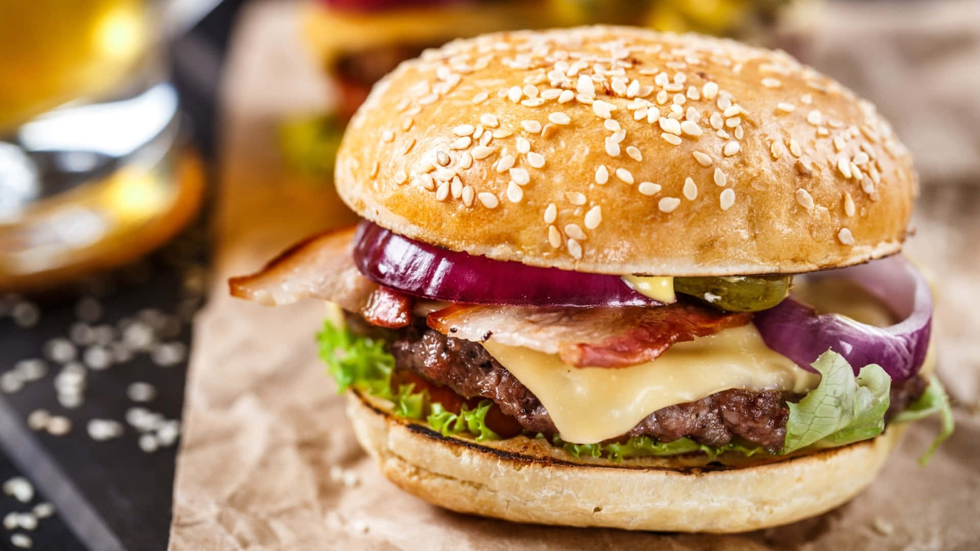 fast food 桌布 壁紙 — 一個多汁的起司漢堡，配上培根、紅洋蔥和生菜，放在芝麻麵包上，擱在皺巴巴的牛皮紙上。