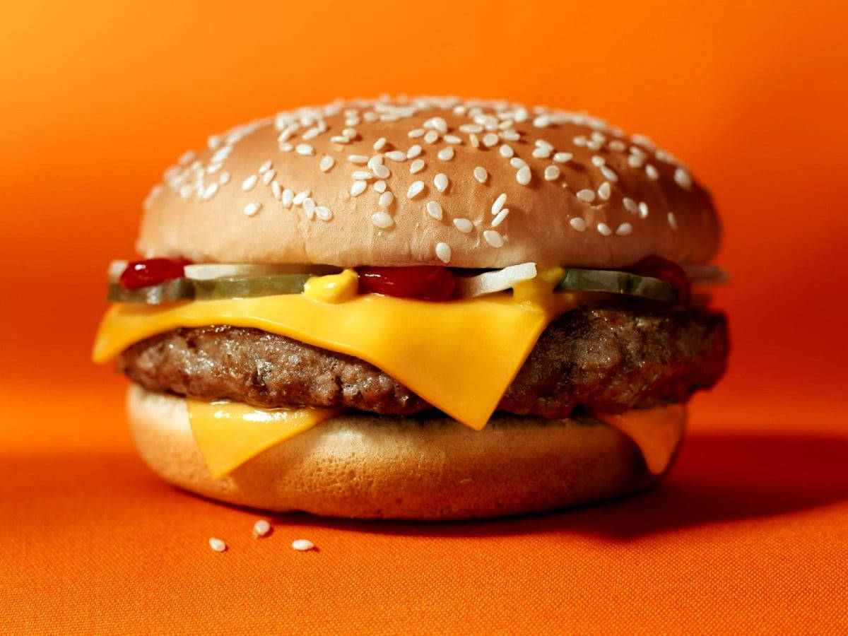 Burger 桌布 壁紙 — 一個多汁的起司漢堡，配芝麻麵包、酸黃瓜和番茄醬，放在鮮豔的橙色背景上。