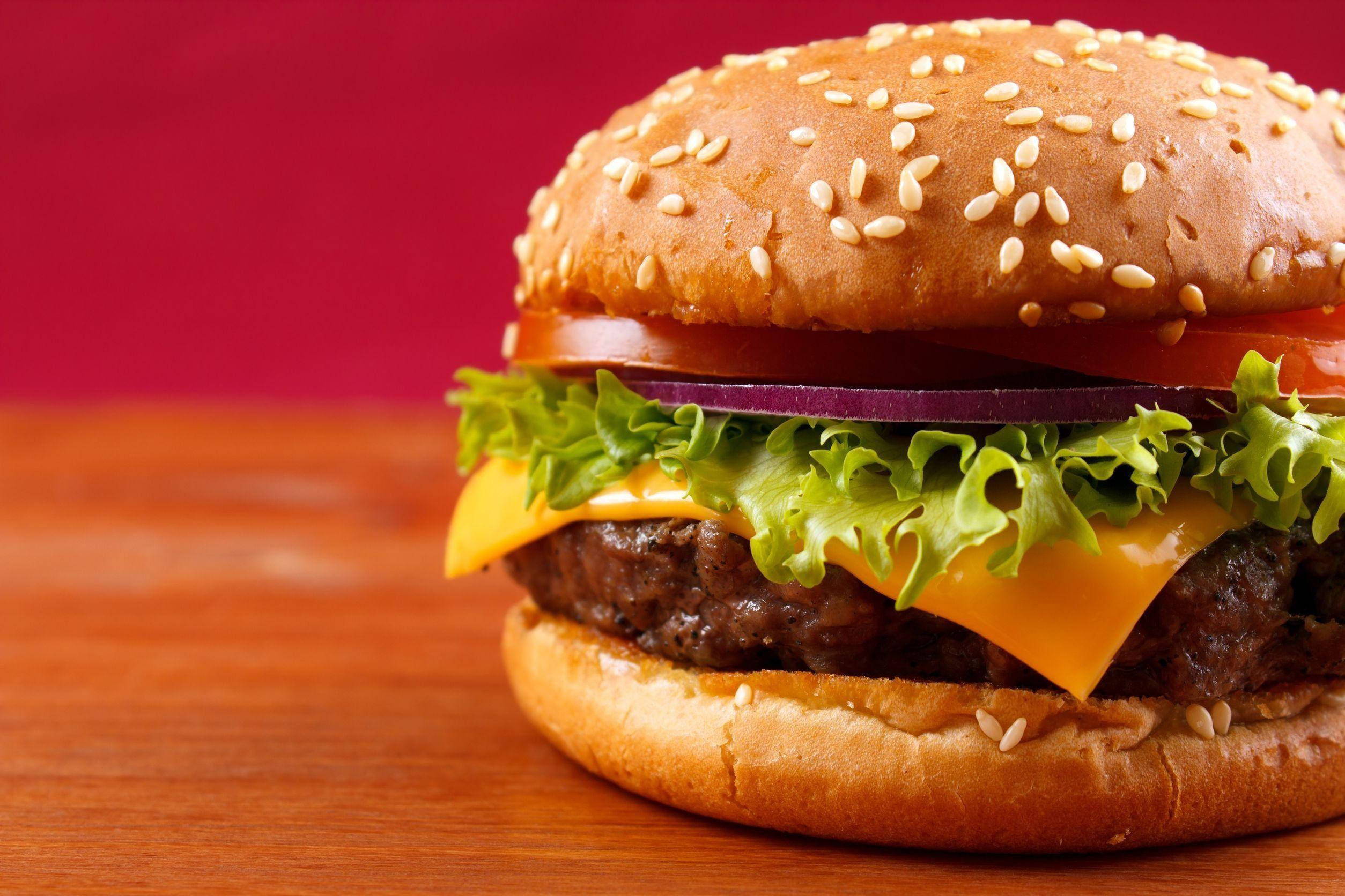 Burger 桌布 壁紙 — 一個多汁的起司漢堡，配芝麻麵包、生菜、番茄和洋蔥，放在木質表面上，背景為深紅色。