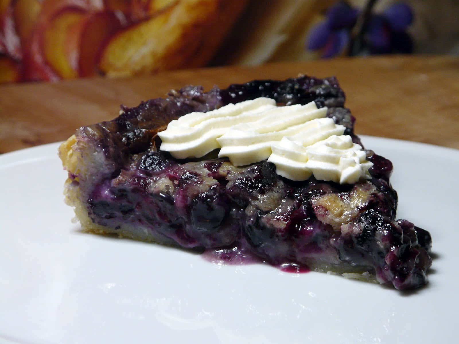 Blueberries Tart 桌布 壁紙 — 一個多汁藍莓派切片的特寫，上面有擠花鮮奶油，放在白色盤子上。