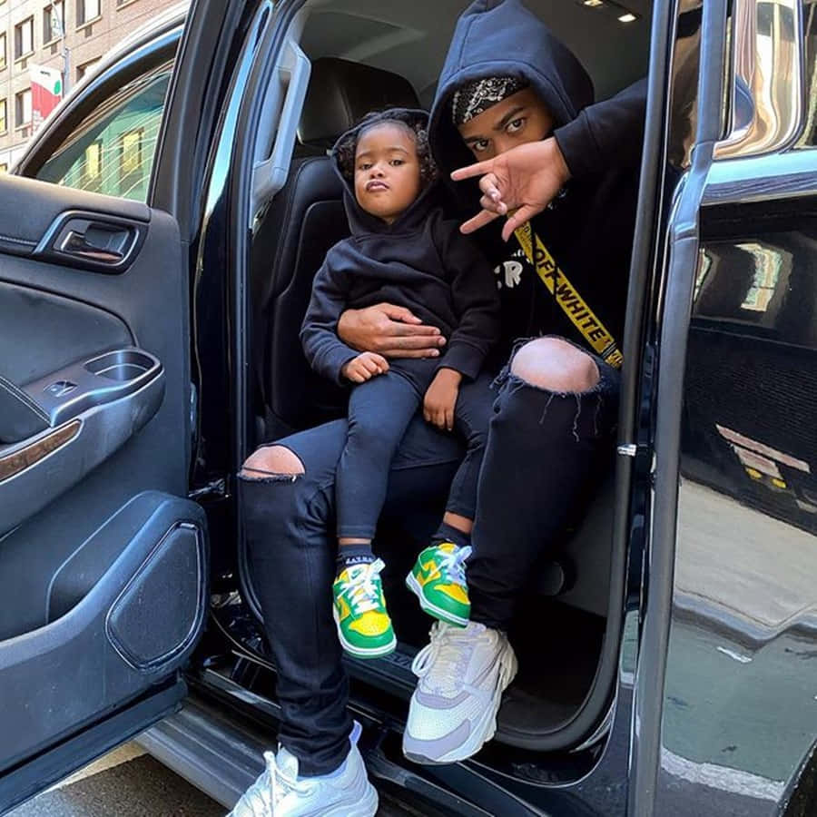 Prettyboyfredo 桌布 壁紙 — 一個大人和一個小孩穿著黑色連帽衫坐在車門邊，擺出各種手勢，腳穿色彩繽紛的運動鞋。