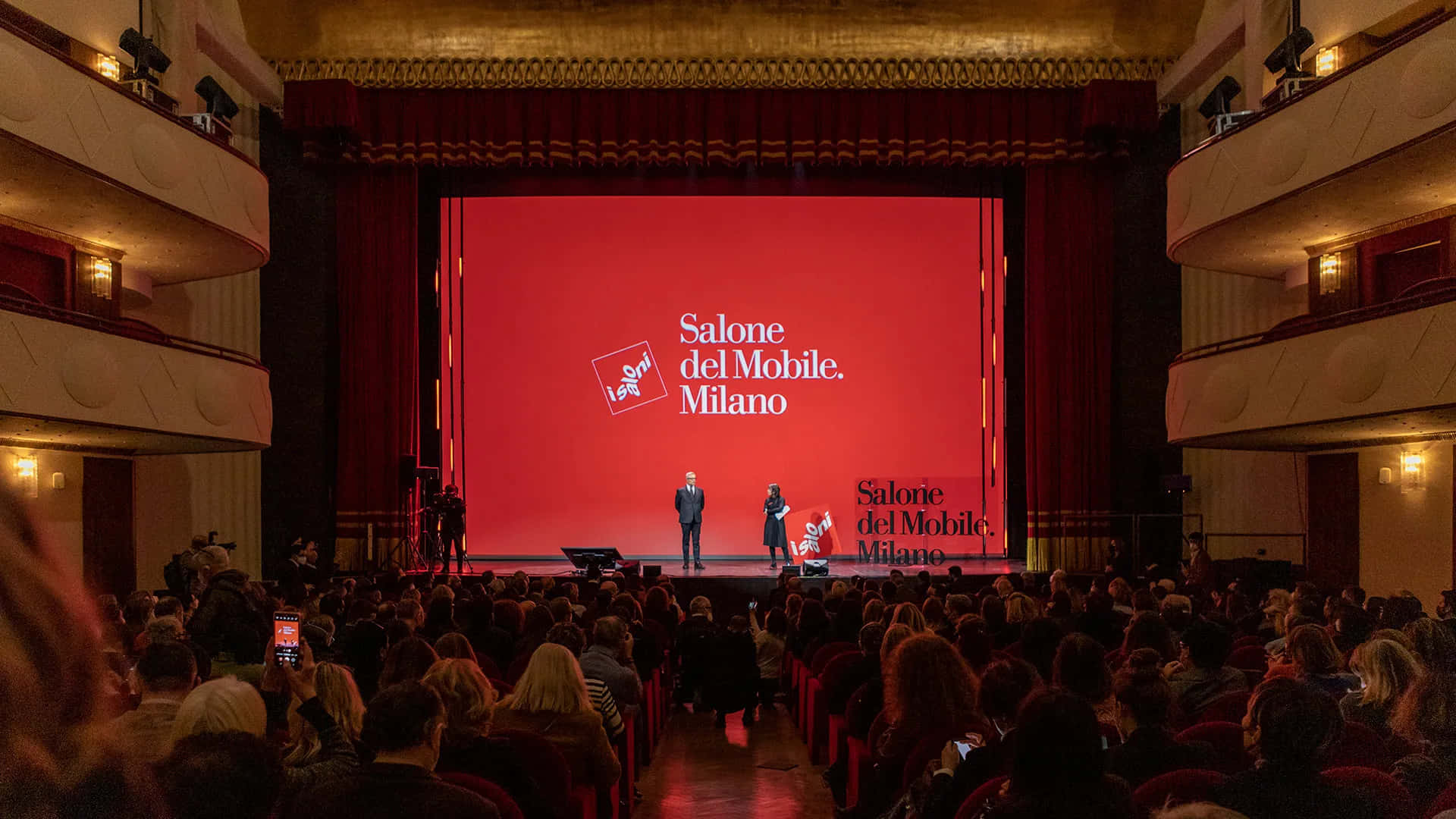 Milan 桌布 壁紙 — 一個大劇院，坐滿觀眾觀看在紅色舞台上展示Salone del Mobile Milano品牌的演示。