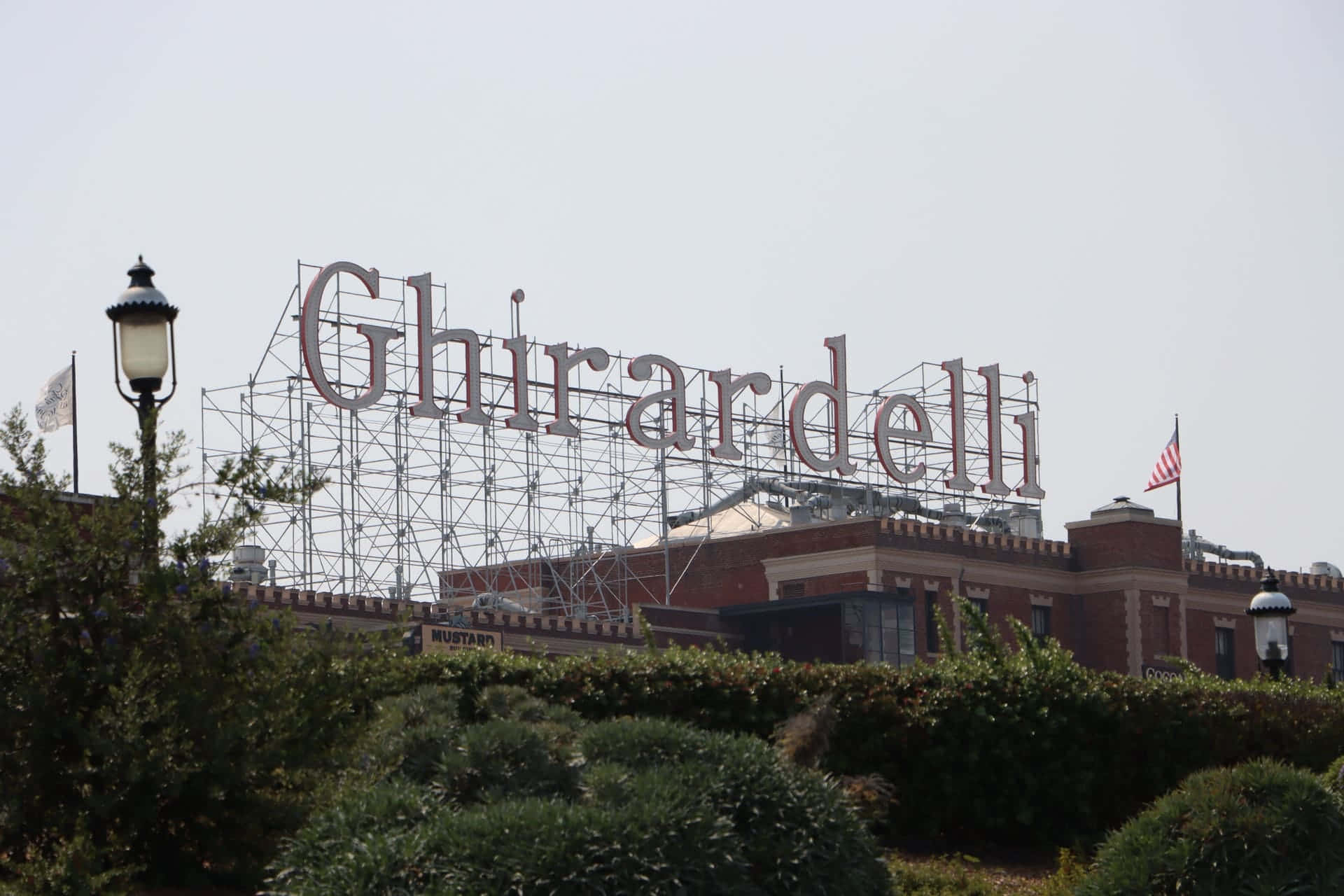 加州 桌布 壁紙 — 一個大型 Ghirardelli 標誌正在紅磚建築上施工，前景有茂密的綠色樹籬。