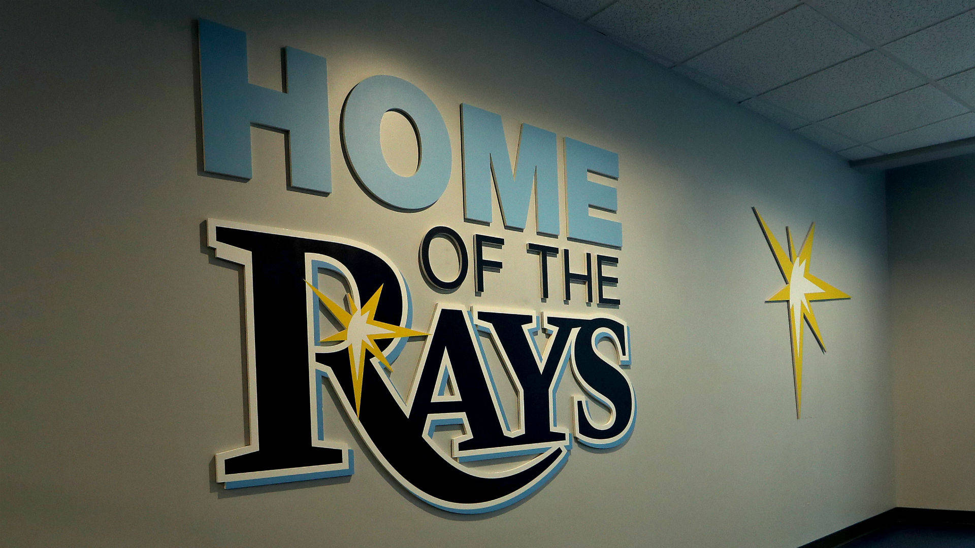 Rays 桌布 壁紙 — 一個大型3D標牌，上面寫著「HOME OF THE RAYS」，並帶有一個黃色星星標誌，安裝在光線昏暗的室內走廊的素牆上。