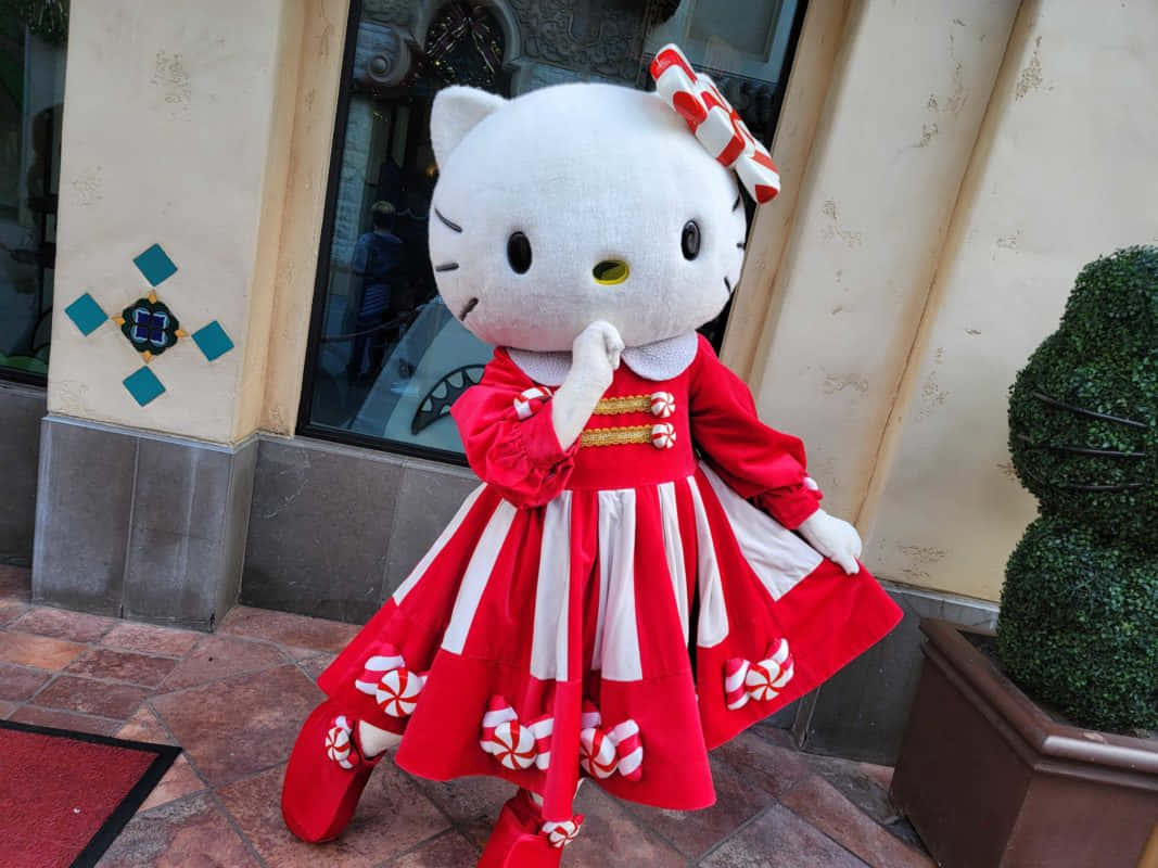 Hello Kitty 桌布 壁紙 — 一個大型Hello Kitty吉祥物穿著節日紅白條紋連衣裙，站在瓷磚走道上，旁邊是一座建築物和盆栽灌木。
