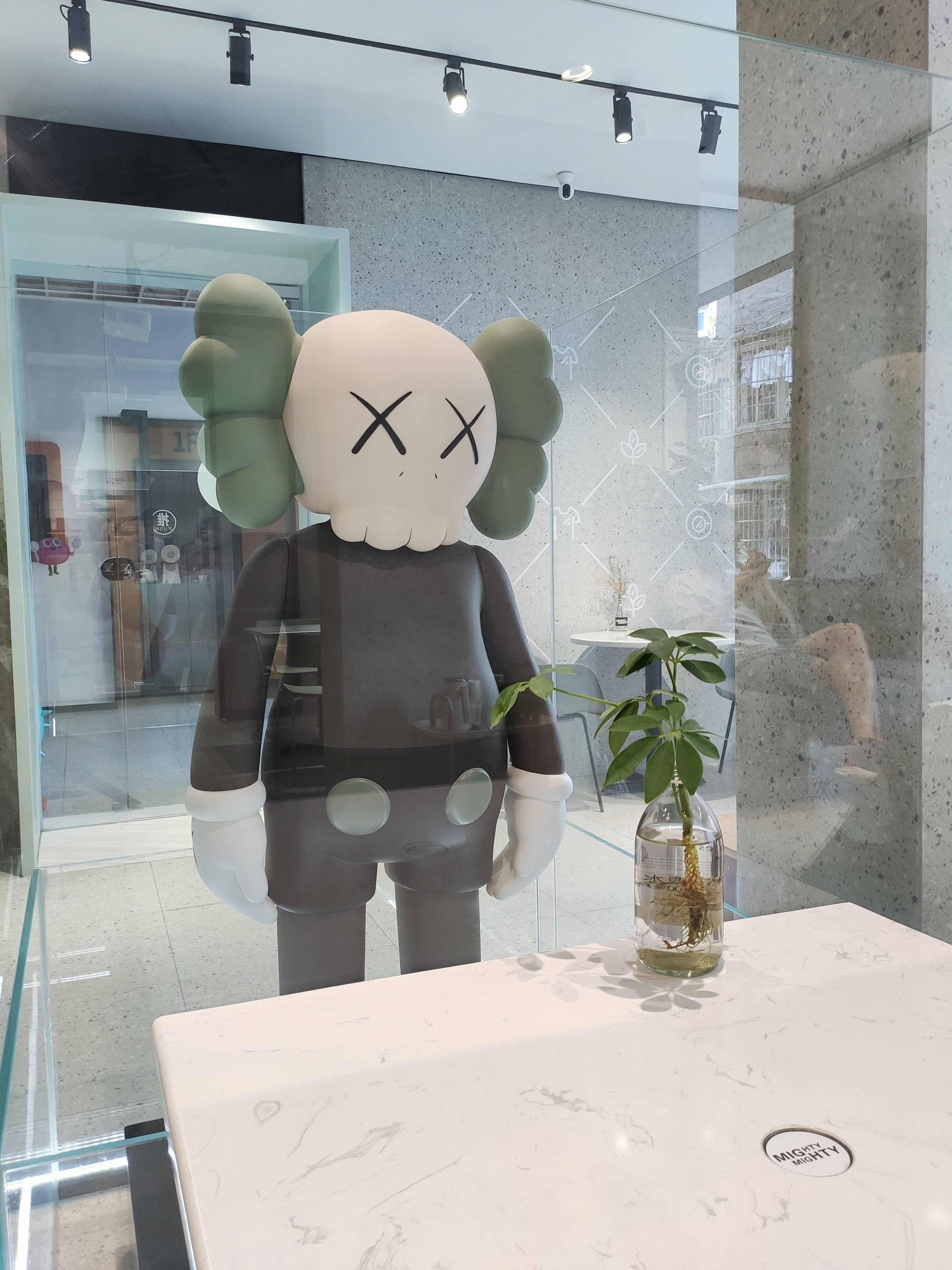 KAWS 桌布 壁紙 — 一個大型KAWS雕塑矗立在現代玻璃牆內部，旁邊是大理石檯面和盆栽植物。