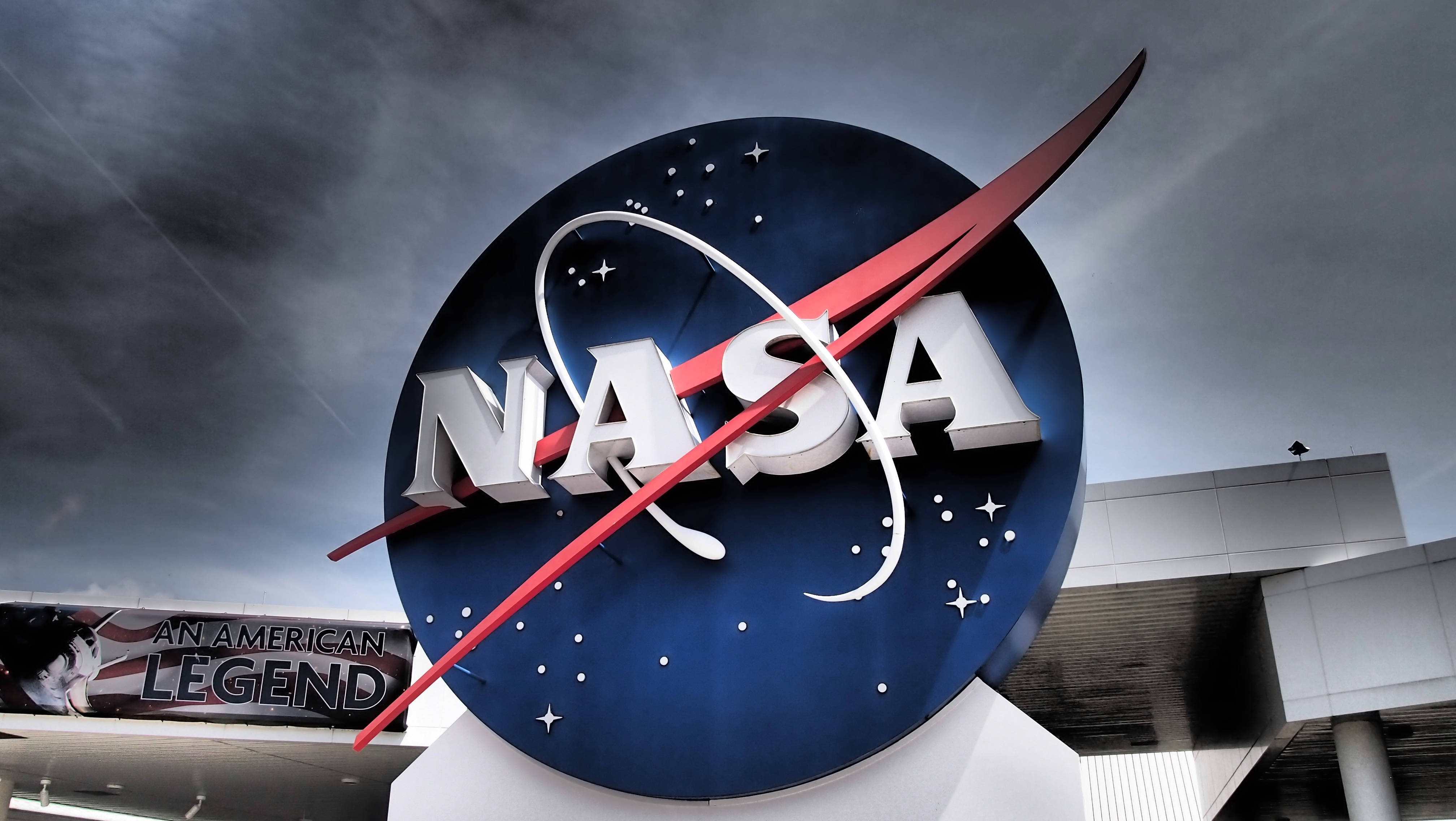 Nasa 桌布 壁紙 — 一個大型NASA標誌雕塑矗立在戲劇性的多雲天空下，背景是一座現代建築。