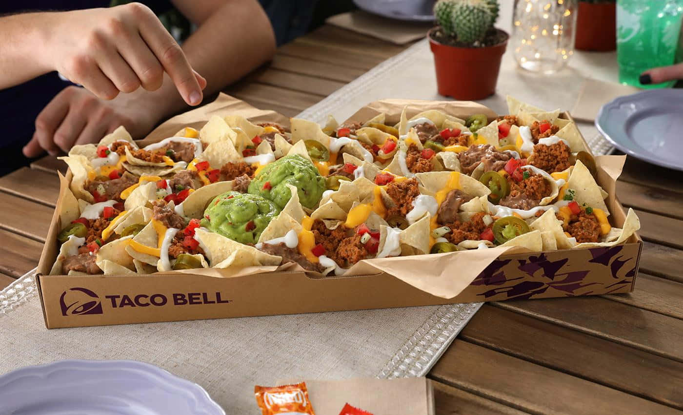 塔可鐘 桌布 壁紙 — 一個大型Taco Bell玉米片盒，裝滿了薯片、起司、酪梨醬和配料，放在木桌上，有手伸進去。
