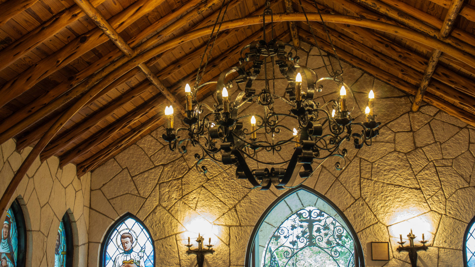 Church 桌布 壁紙 — 一個大型吊燈懸掛在質樸的木樑天花板上，下方是帶有彩繪玻璃窗的石牆。