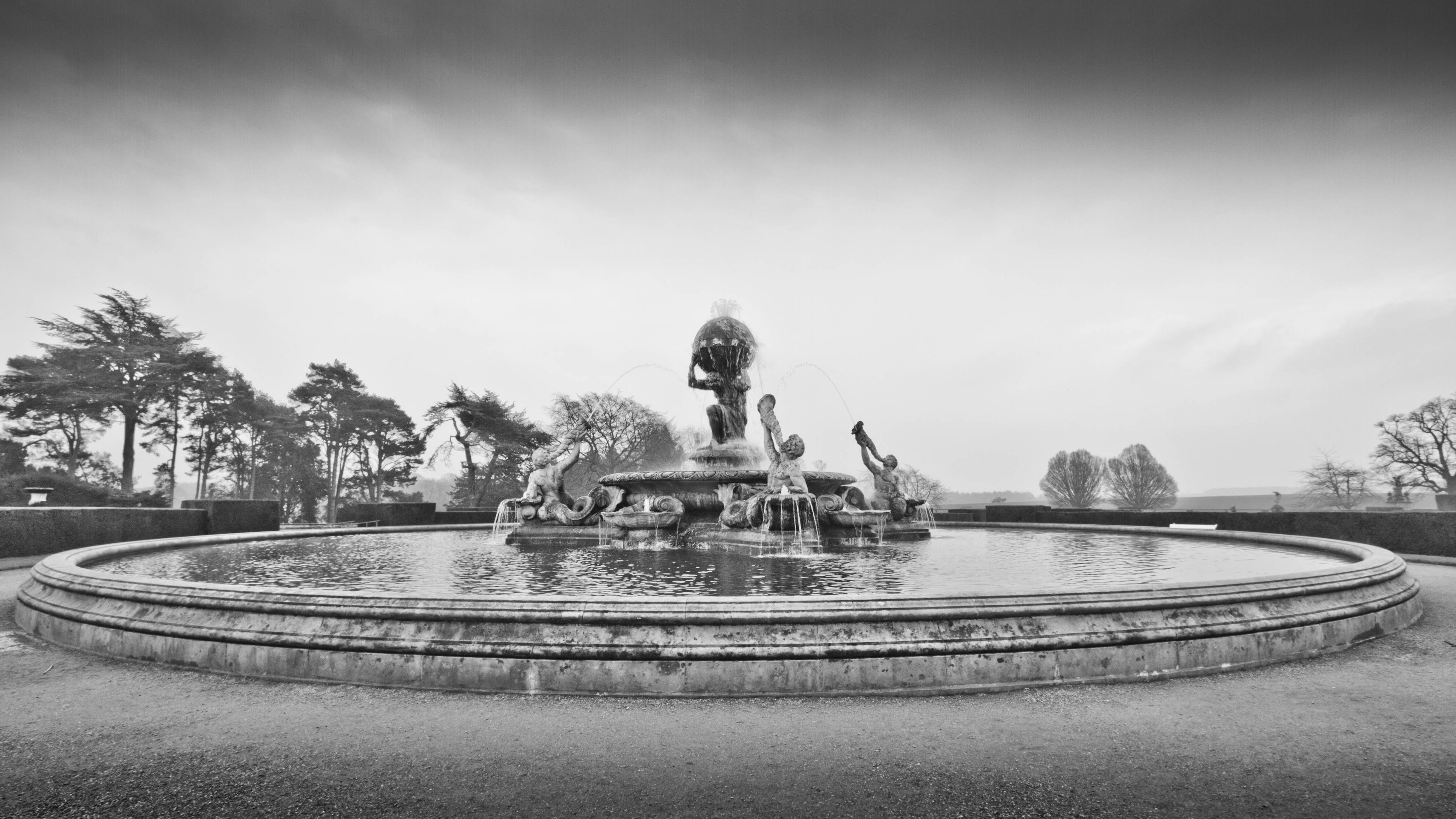 Monochrome 桌布 壁紙 — 一個大型單色噴泉，帶有雕塑人物和層疊流水，坐落於陰天下的正式花園中。