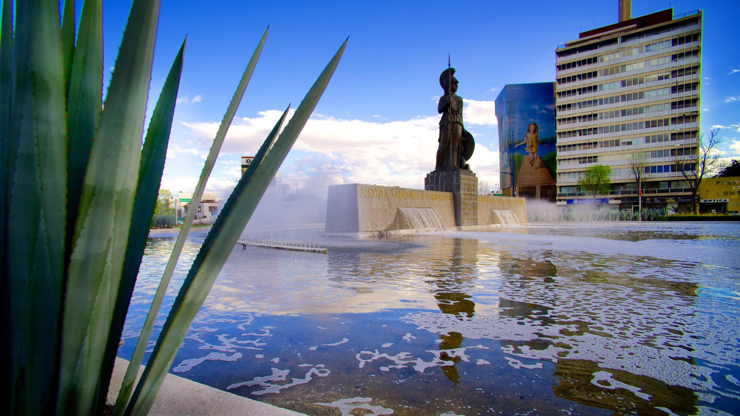 Guadalajara 桌布 壁紙 — 一個大型噴泉，中央有一座戰士青銅雕像，矗立在陽光明媚的城市廣場上，周圍環繞著高大的龍舌蘭葉和現代建築。