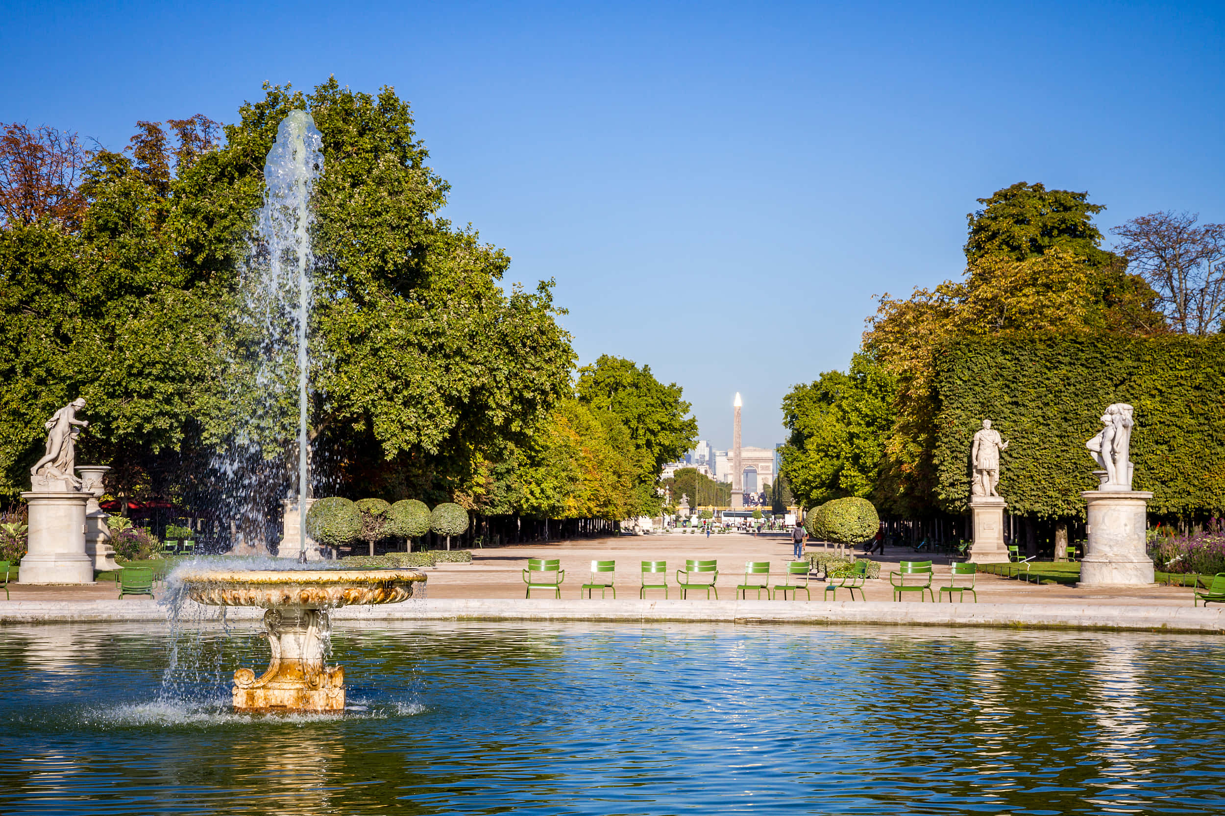 公園 桌布 壁紙 — 一個大型噴泉在陽光明媚的公園中噴水，公園兩旁有雕像、綠色樹籬和遠處的城市天際線。