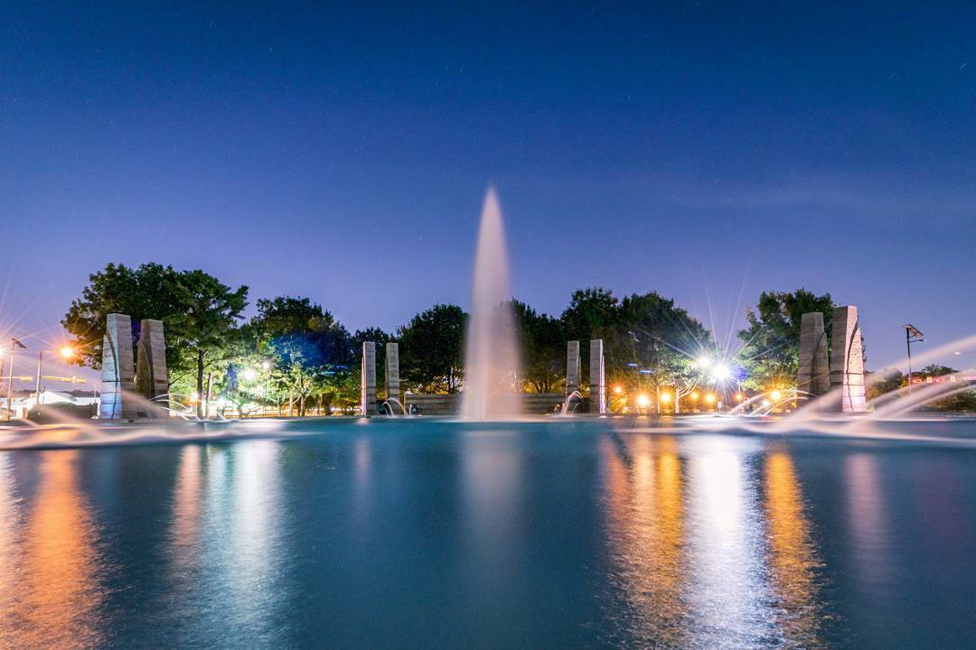 HD Water 桌布 壁紙 — 一個大型噴泉將水噴向暮色天空，城市燈光映照在平靜的水面上，四周環繞樹木。