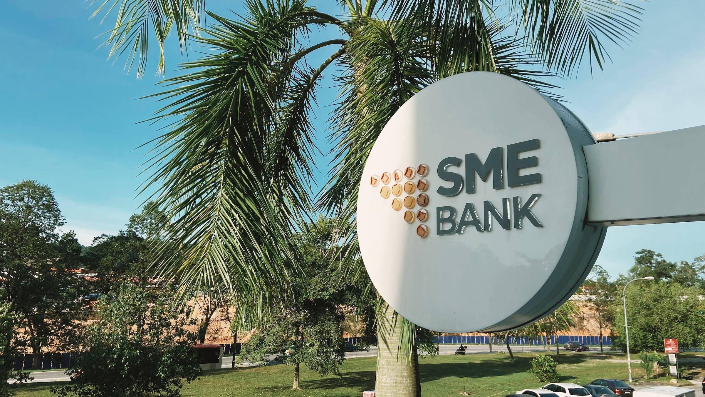 設計 桌布 壁紙 — 一個大型圓形SME Bank標誌在晴朗的藍天下格外醒目，周圍環繞著茂密的綠色棕櫚葉和樹木。