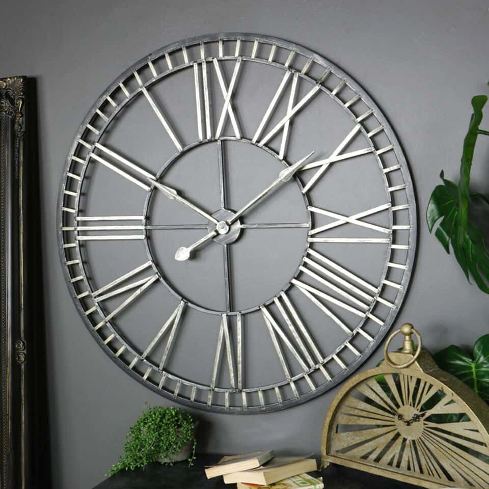 Wall Clock 桌布 壁紙 — 一個大型工業風格的掛牆鐘，帶有羅馬數字，掛在灰色牆上，旁邊有綠色植物和復古裝飾。