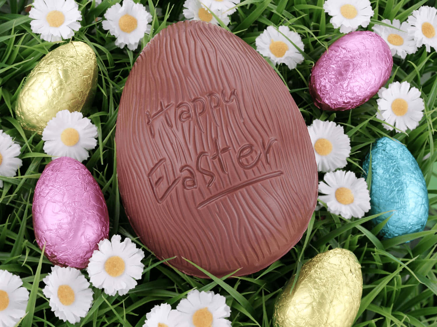 復活節 桌布 壁紙 — 一個大型巧克力復活蛋，上面壓印了「Happy Easter」，放在綠色草地上，周圍有錫箔紙包裝的蛋和雛菊。