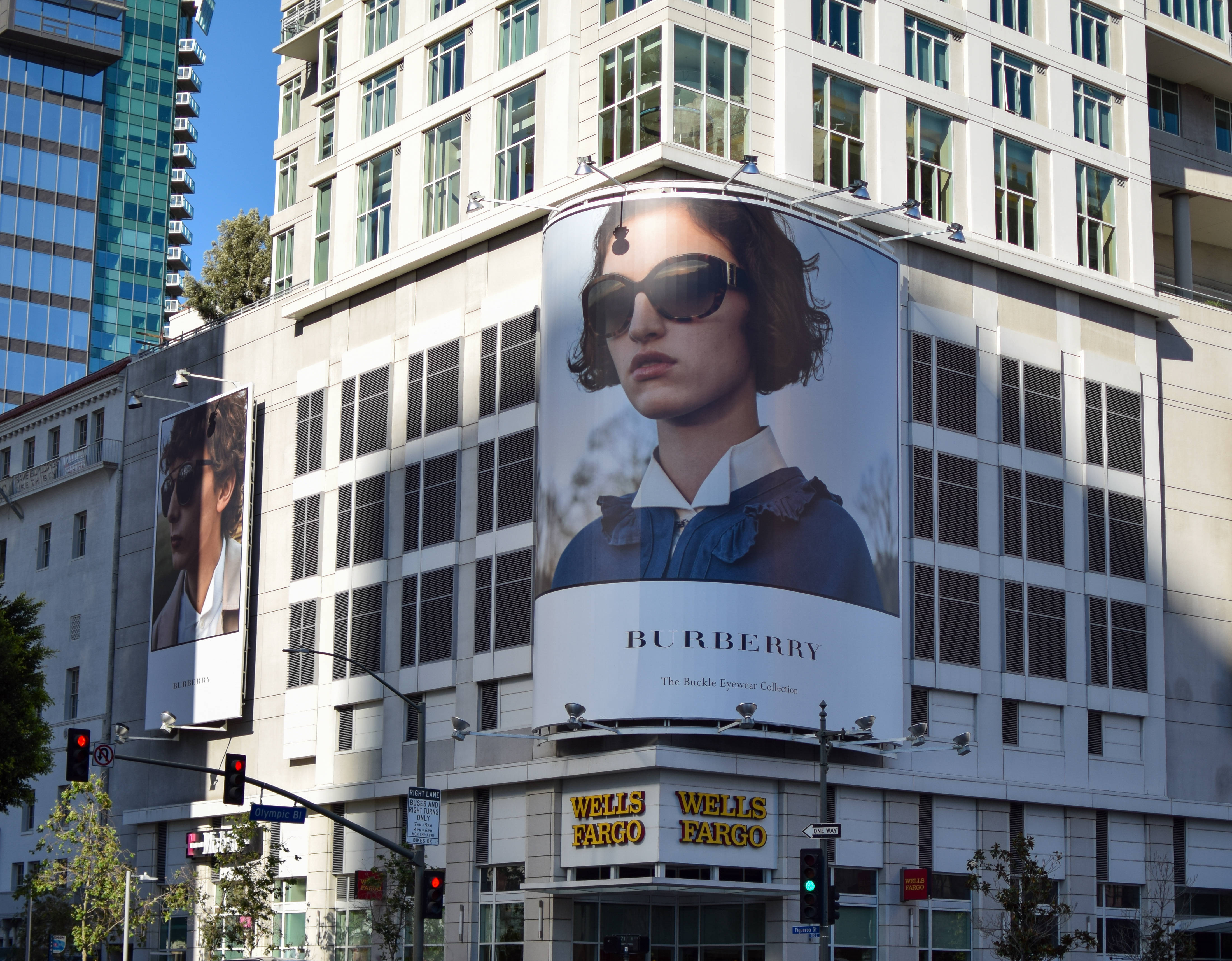 Burberry 桌布 壁紙 — 一個大型彎曲的Burberry廣告牌，上面有一位戴太陽眼鏡的女性，在晴朗藍天下主導著現代城市建築的角落。