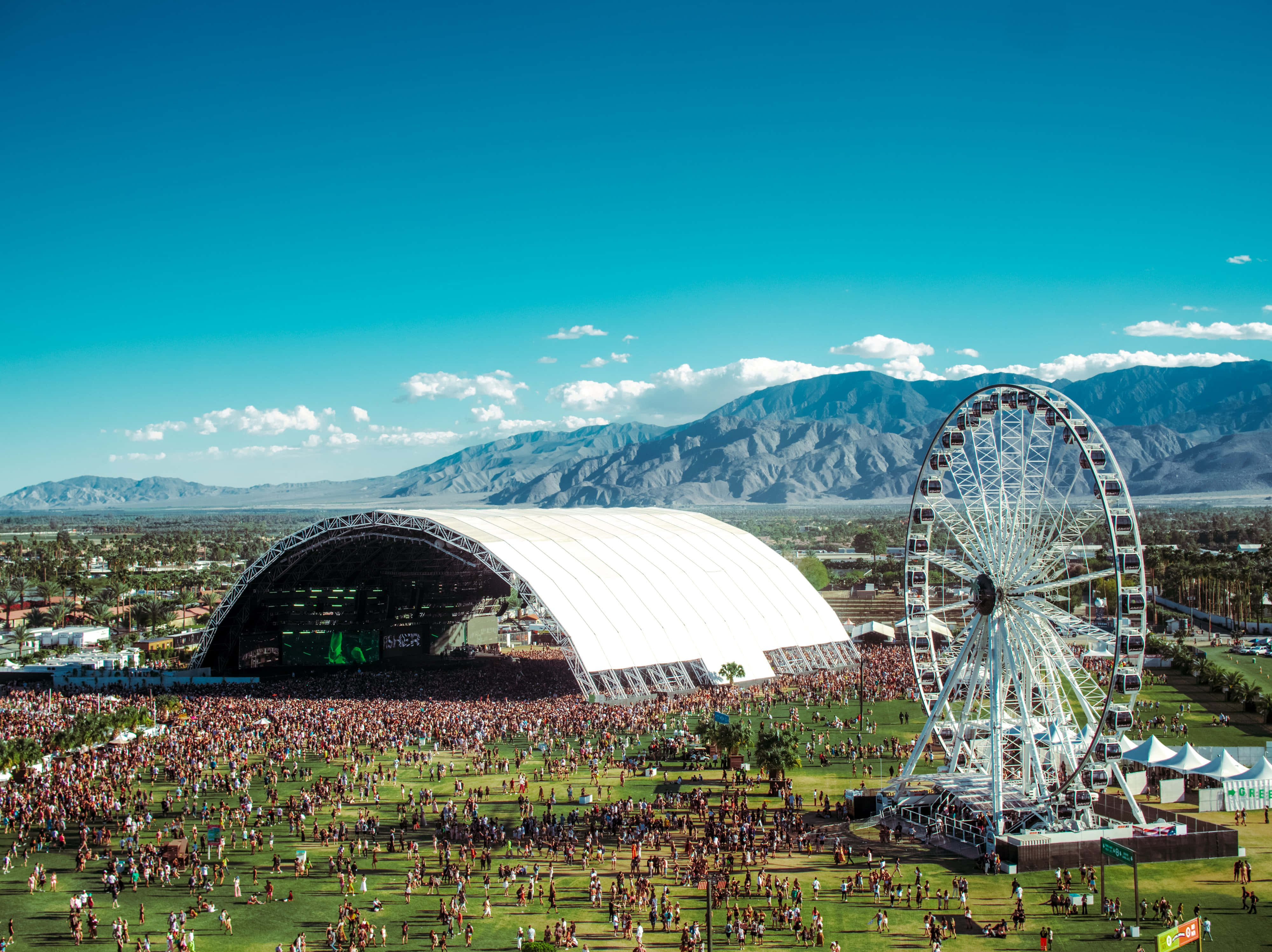 Coachella 桌布 壁紙 — 一個大型戶外音樂節，設有巨大的白色圓頂舞台和摩天輪，背景是明亮的藍天和遠處的山脈。