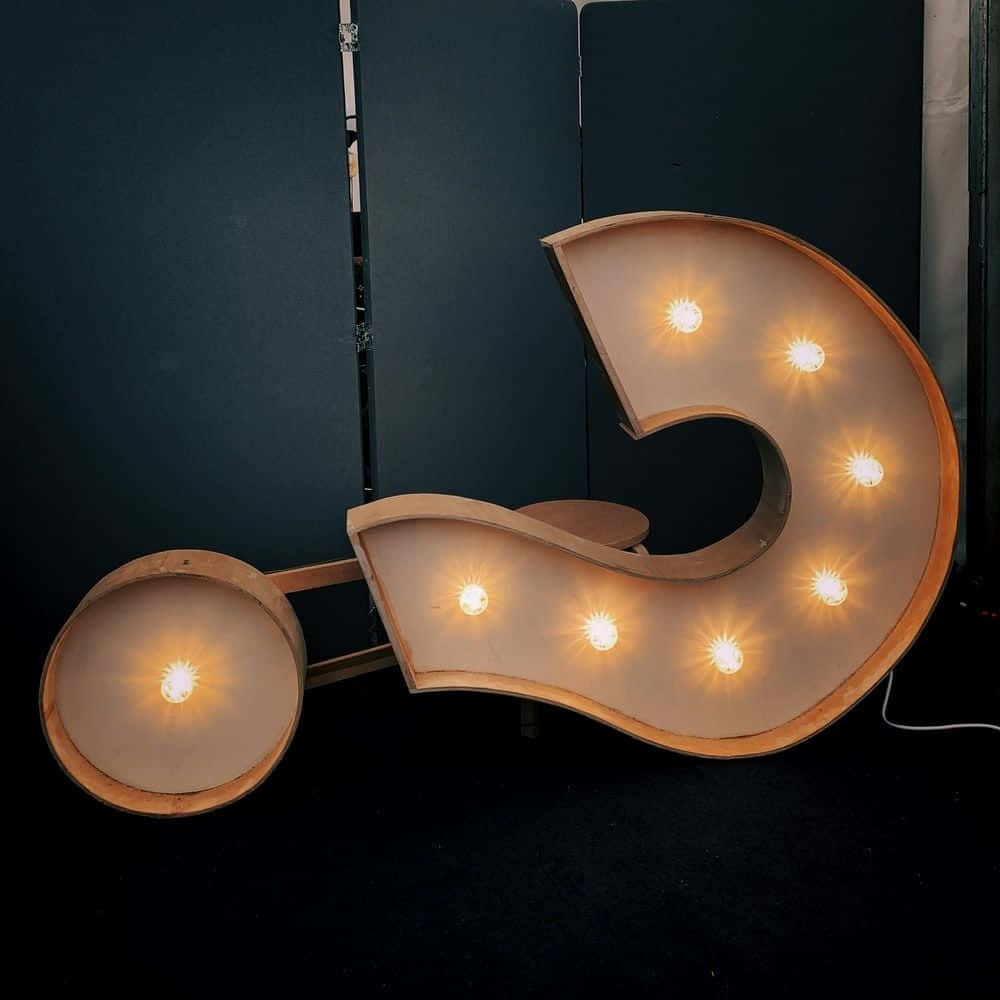 Question Mark 桌布 壁紙 — 一個大型木製問號標誌，帶有發光的燈泡，立在黑暗背景前，投射出溫暖的光芒。