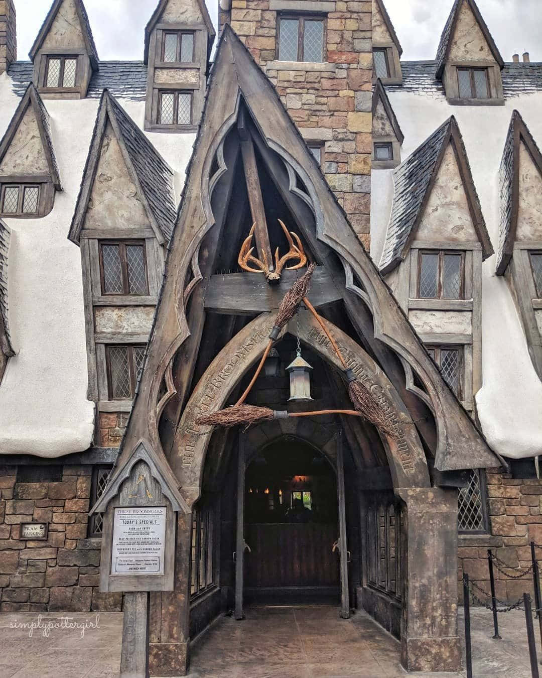 Hogsmeade 桌布 壁紙 — 一個大型木製拱門，裝飾著掃帚和鹿角，框住了一座質樸的石頭和灰泥建築的入口，天空多雲。
