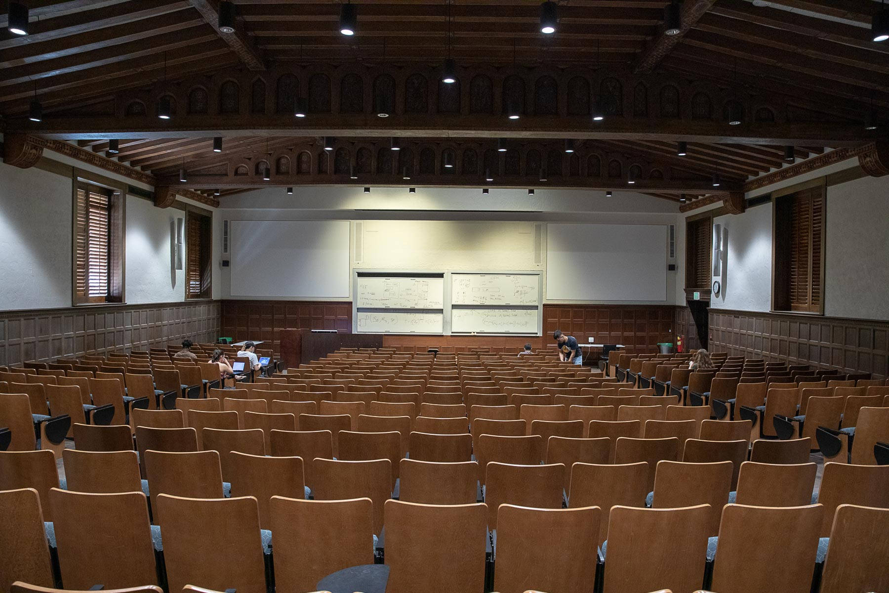 ucla 桌布 壁紙 — 一個大型木製演講廳，在溫暖的頂燈下，前排白板附近有幾排空椅和幾名學生。