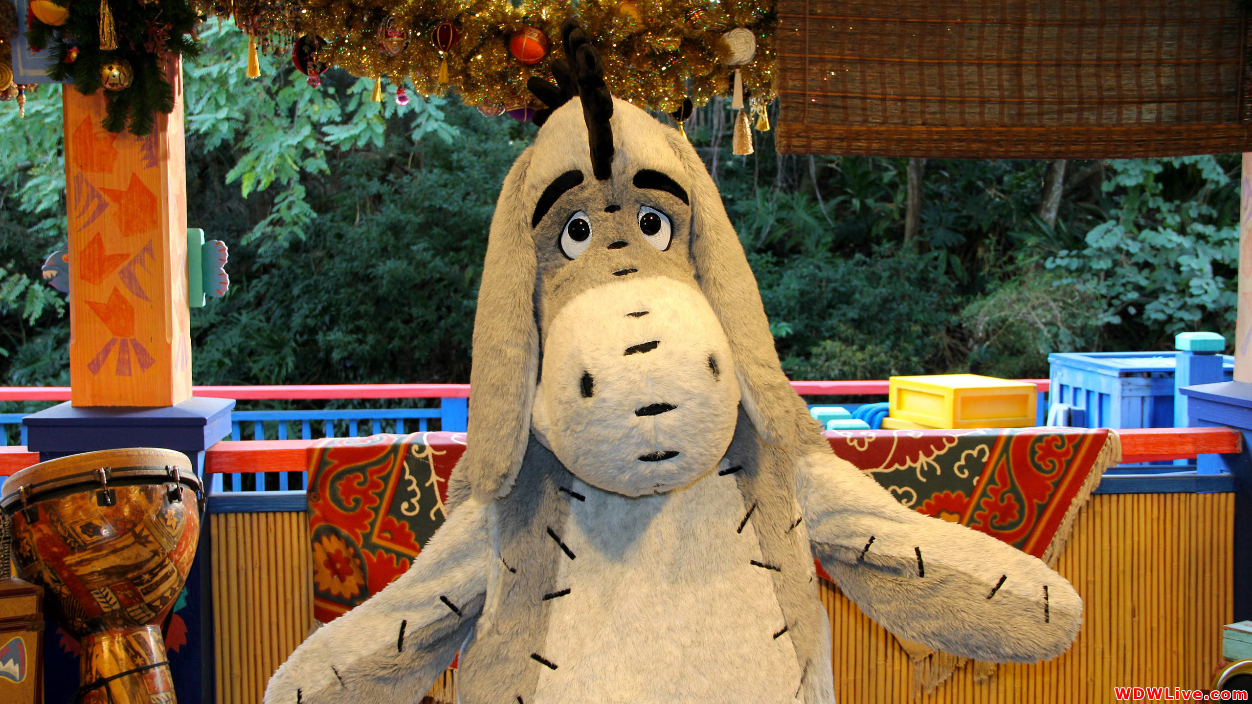 Eeyore 桌布 壁紙 — 一個大型毛絨伊唷公仔站在色彩繽紛的裝飾門廊上，周圍環繞著節日裝飾和叢林主題道具。