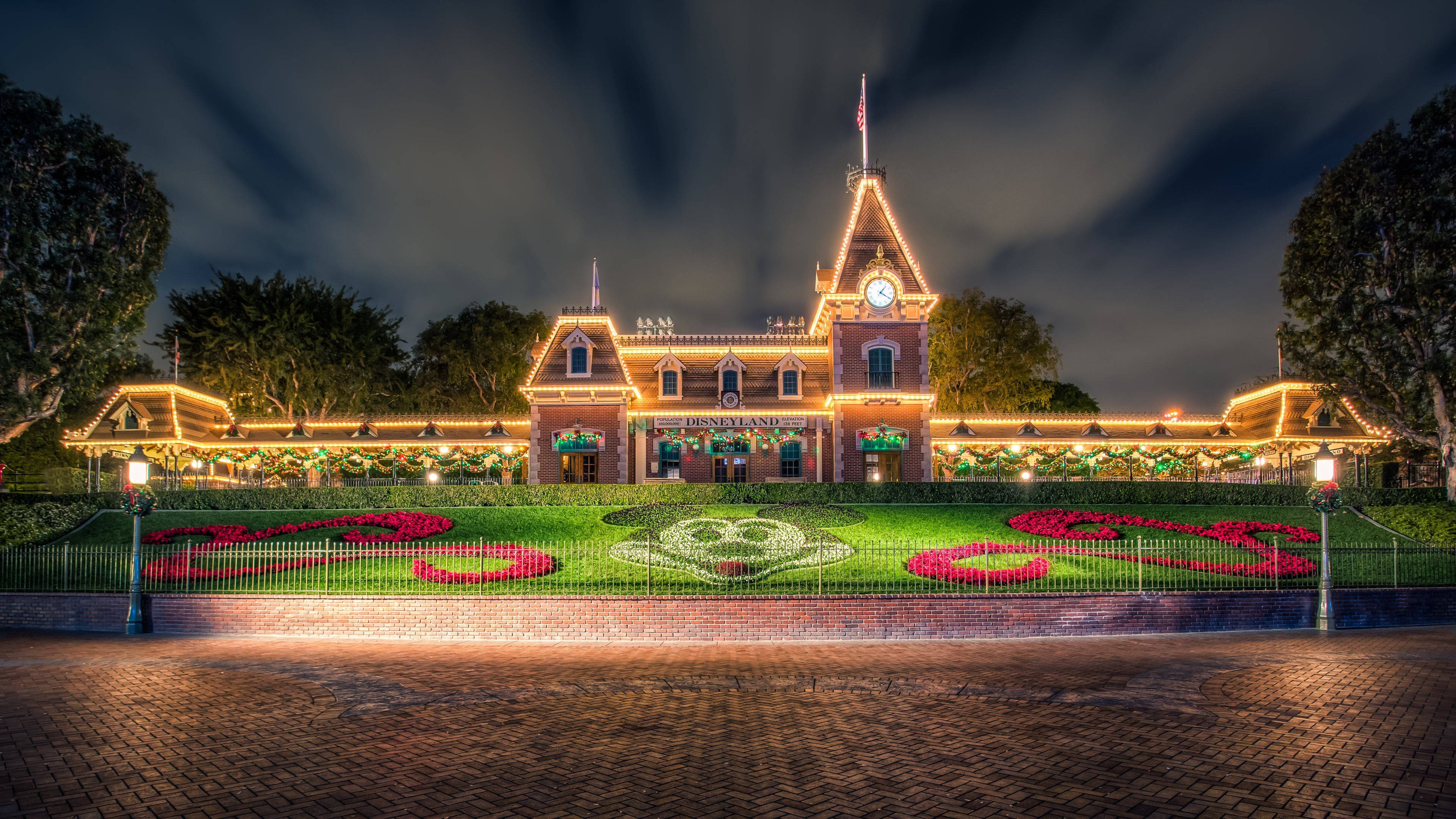 Disneyland 桌布 壁紙 — 一個大型燈光火車站夜晚裝飾著節日燈光和前面充滿活力的花卉米奇老鼠設計。