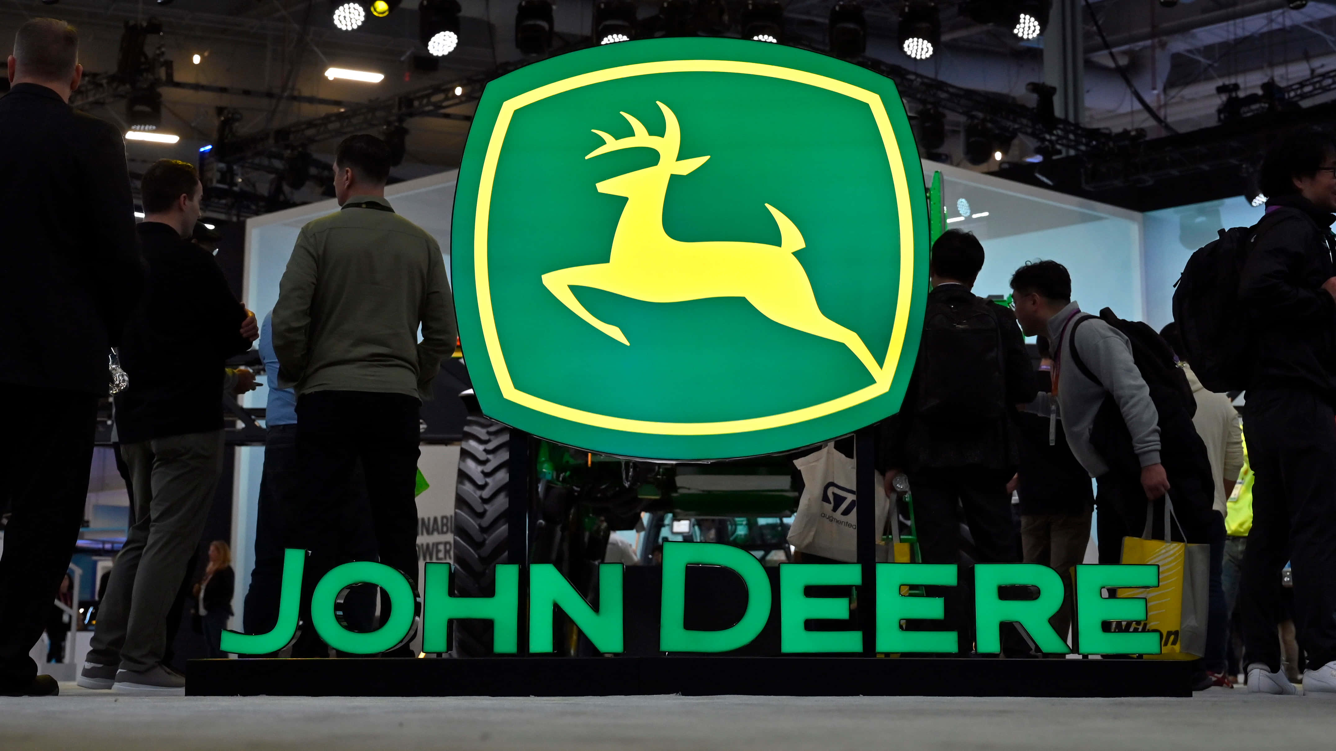 約翰迪爾 桌布 壁紙 — 一個大型發光的John Deere標誌，帶有一隻跳躍的鹿，在繁忙的室內展覽廳的貿易展上醒目地矗立，周圍有與會者。