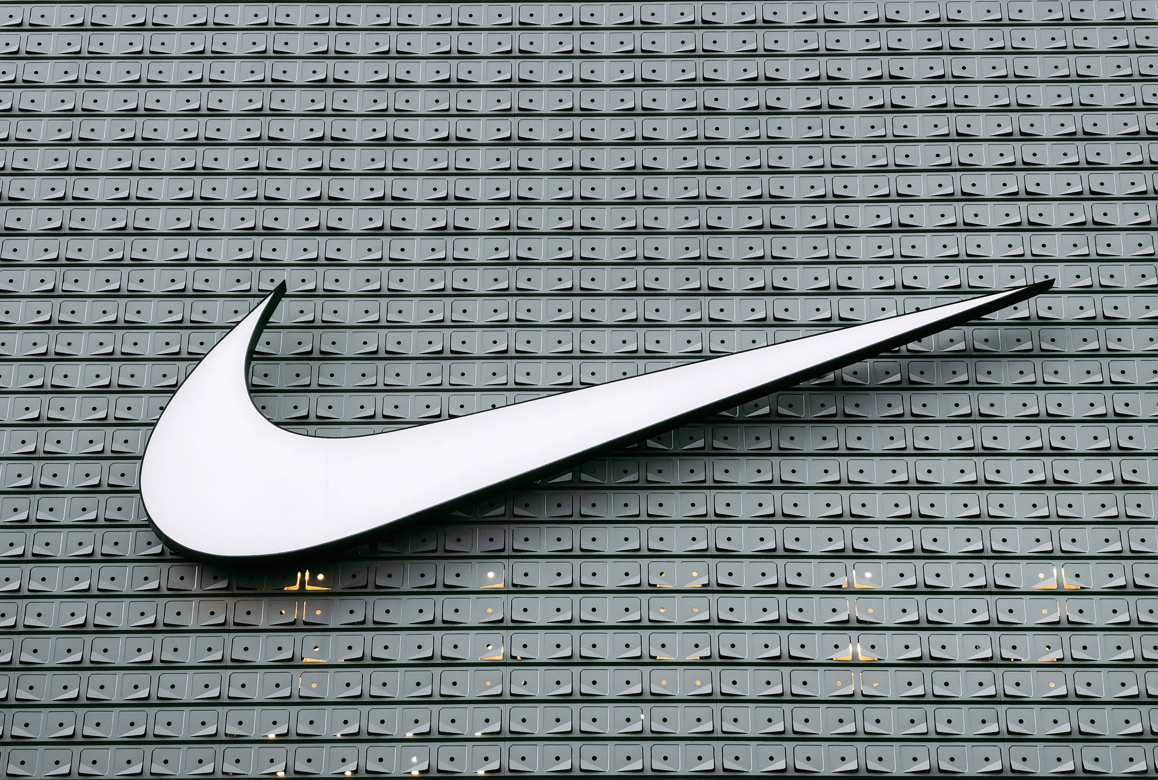 Swoosh 桌布 壁紙 — 一個大型白色Nike swoosh標誌安裝在帶有重複方形圖案的灰色紋理牆上。