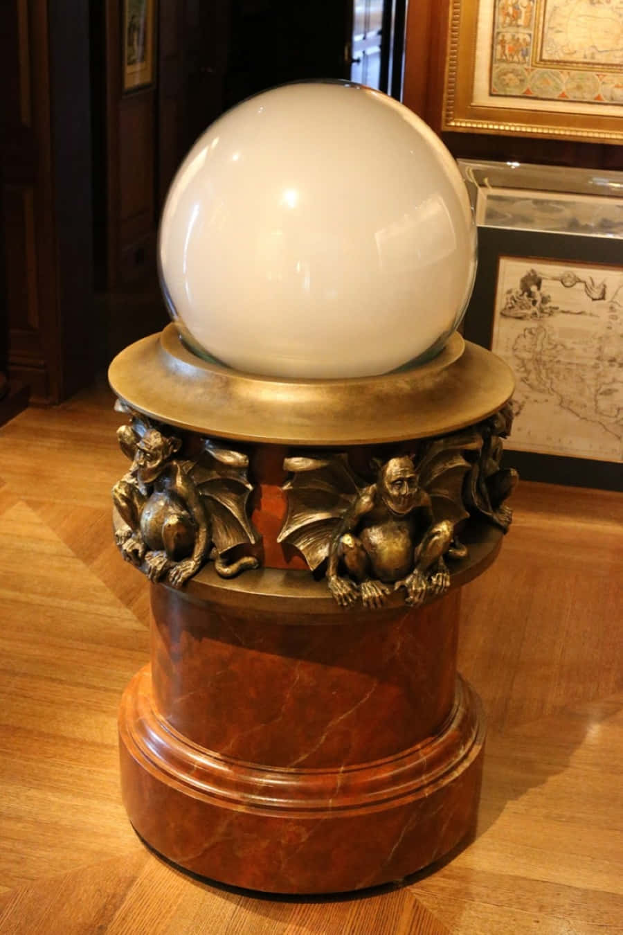 Crystal Ball 桌布 壁紙 — 一個大型白色玻璃球體放置在一個飾有石像鬼圖案的青銅底座上，底座置於一個拋光紅色大理石基座上，位於木質房間內。