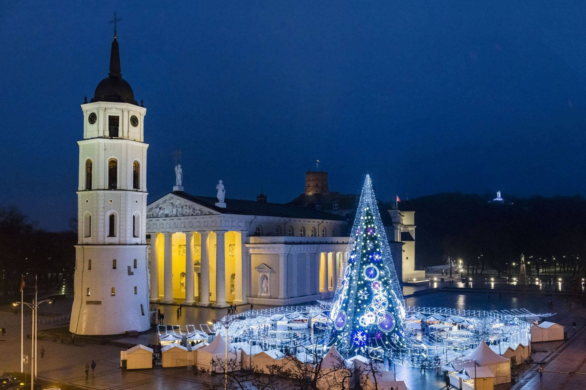 Vilnius 桌布 壁紙 — 一個大型的、亮燈的聖誕樹矗立在雪地廣場上，旁邊是一座歷史悠久的白色大教堂，正值黃昏時分。