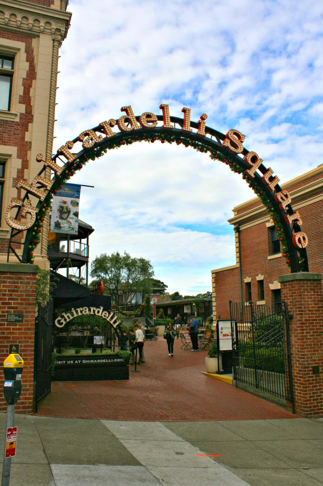 加州 桌布 壁紙 — 一個大型磚拱門，上面有金色字樣的「Ghirardelli Square」，框住了一條在部分多雲天空下繁忙的行人步道。