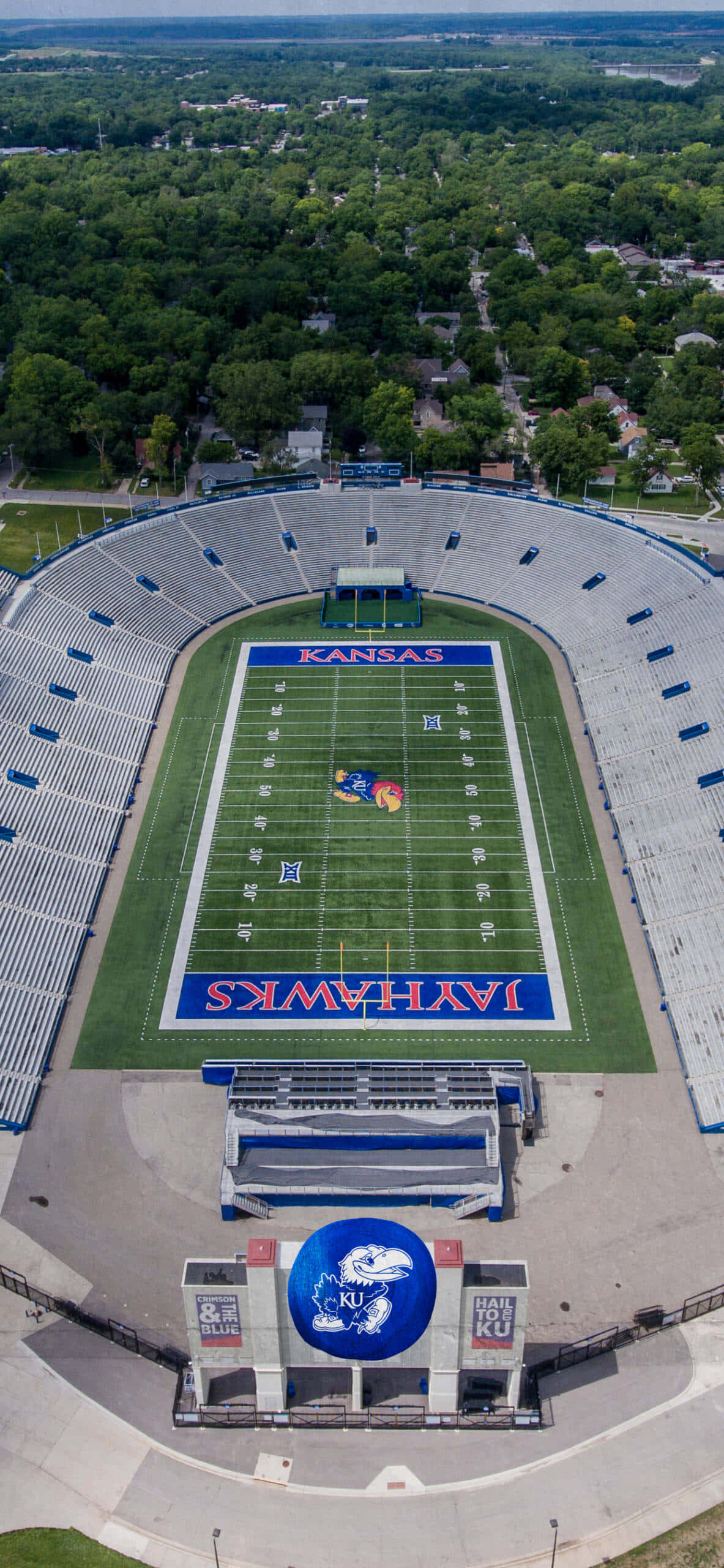Stadium 桌布 壁紙 — 一個大型空蕩蕩的足球場的鳥瞰圖，綠色球場上塗有「KANSAS JAYHAWKS」字樣，周圍環繞著茂密的綠色樹木。