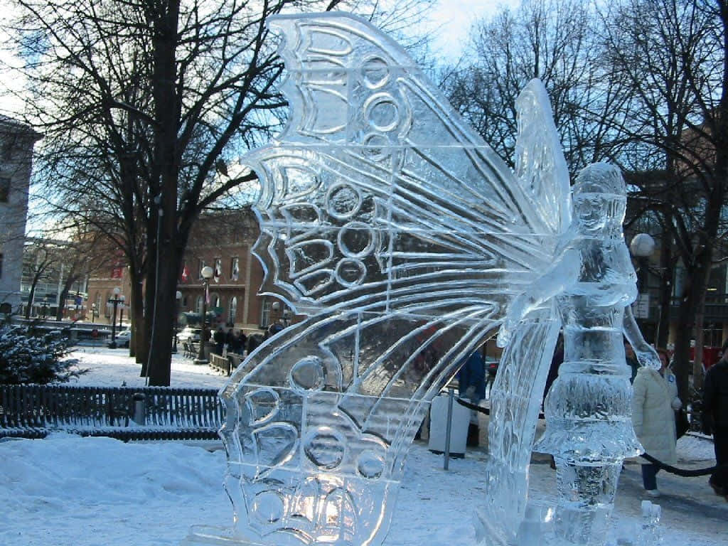 Ice Sculpture 桌布 壁紙 — 一個大型精緻的蝴蝶冰雕，翅膀細節豐富，矗立在晴朗藍天下白雪覆蓋的城市公園中。
