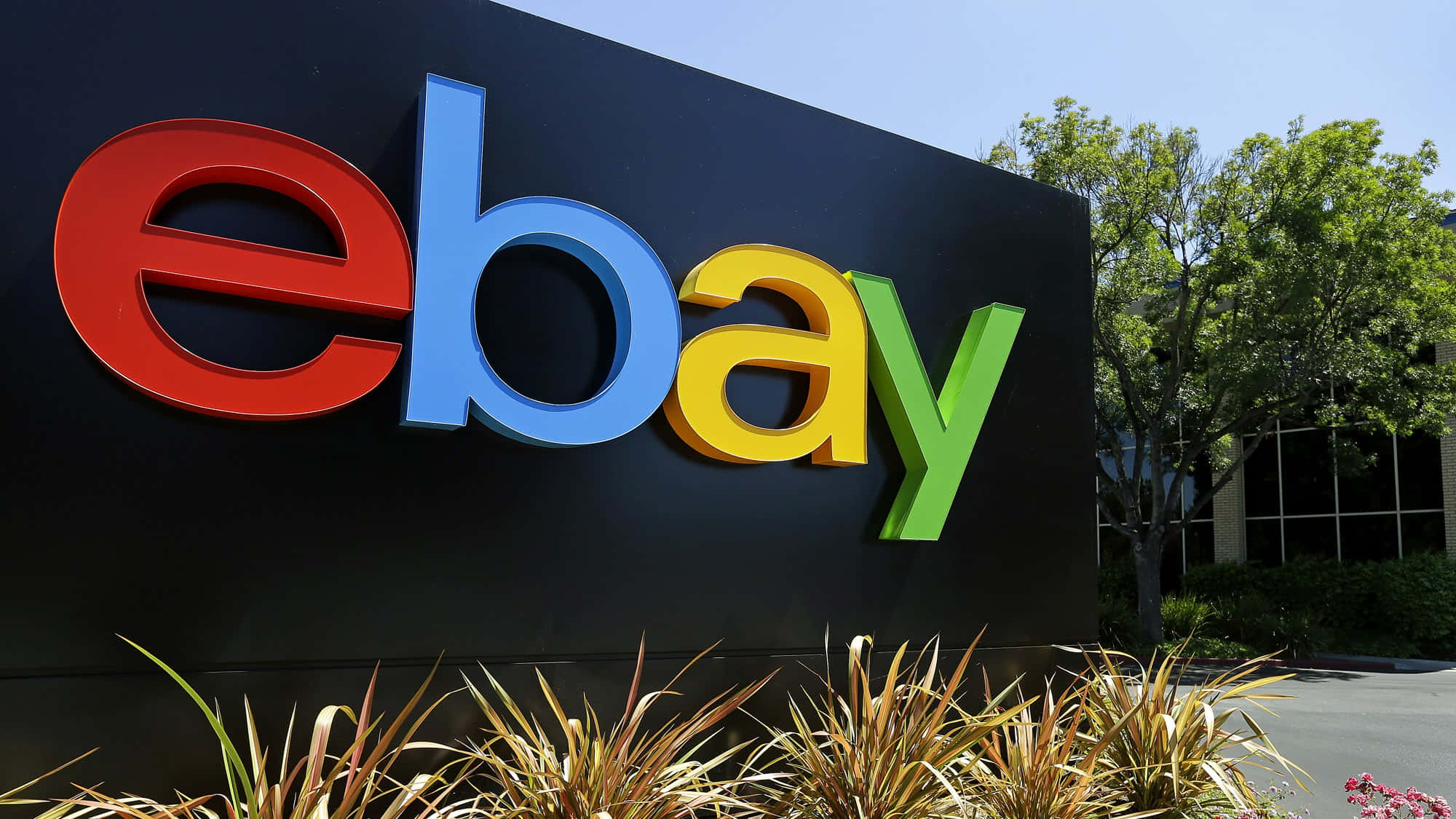 eBay 桌布 壁紙 — 一個大型色彩繽紛的eBay標誌招牌矗立在戶外，藍天白雲下，前景有綠樹和觀賞植物。