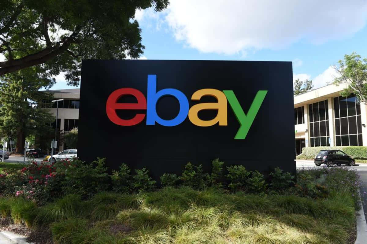 標誌 桌布 壁紙 — 一個大型色彩繽紛的eBay標誌牌，醒目地矗立在一棟現代辦公大樓前，背景是晴朗的藍天和零星的白雲。