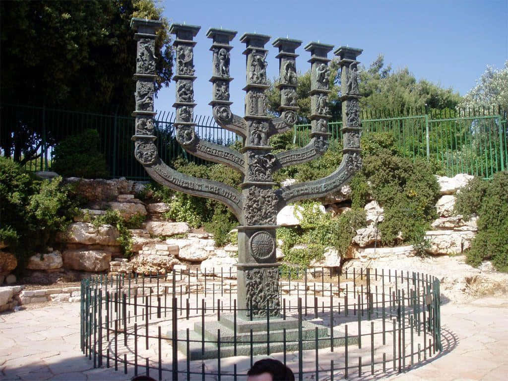 Israel 桌布 壁紙 — 一個大型華麗的青銅 menorah 雕塑矗立在陽光普照的花園中，周圍環繞著綠植和石牆。