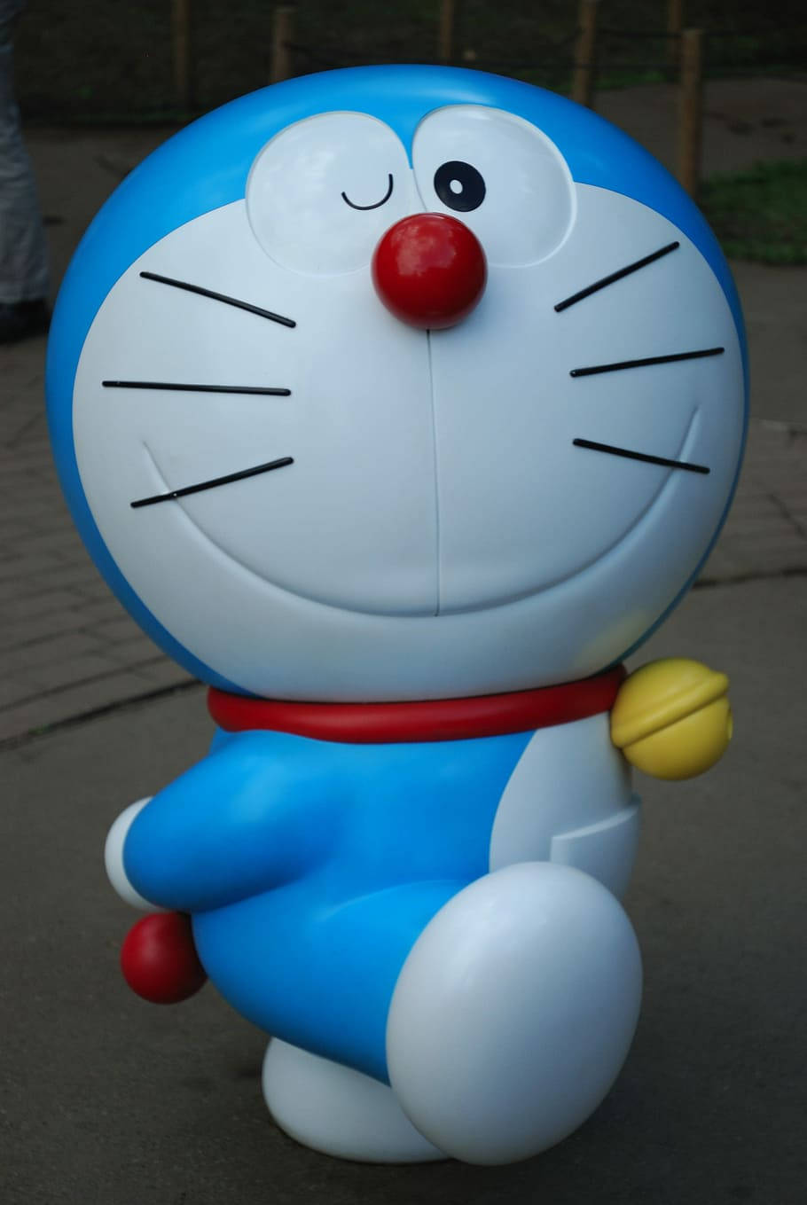 Doraemon 桌布 壁紙 — 一個大型藍色機器貓雕像，有紅色鼻子和黃色鈴鐺，微笑地坐在鋪砌的小路上。