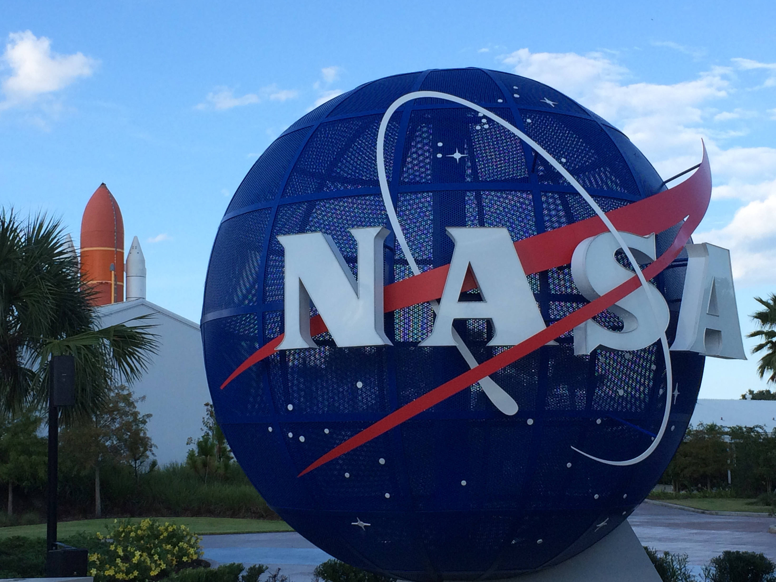 NASA 動態圖片 桌布 壁紙 — 一個大型藍色網狀NASA標誌球體，帶有紅色和白色弧線，矗立在戶外，天空多雲，背景中可見一枚火箭。