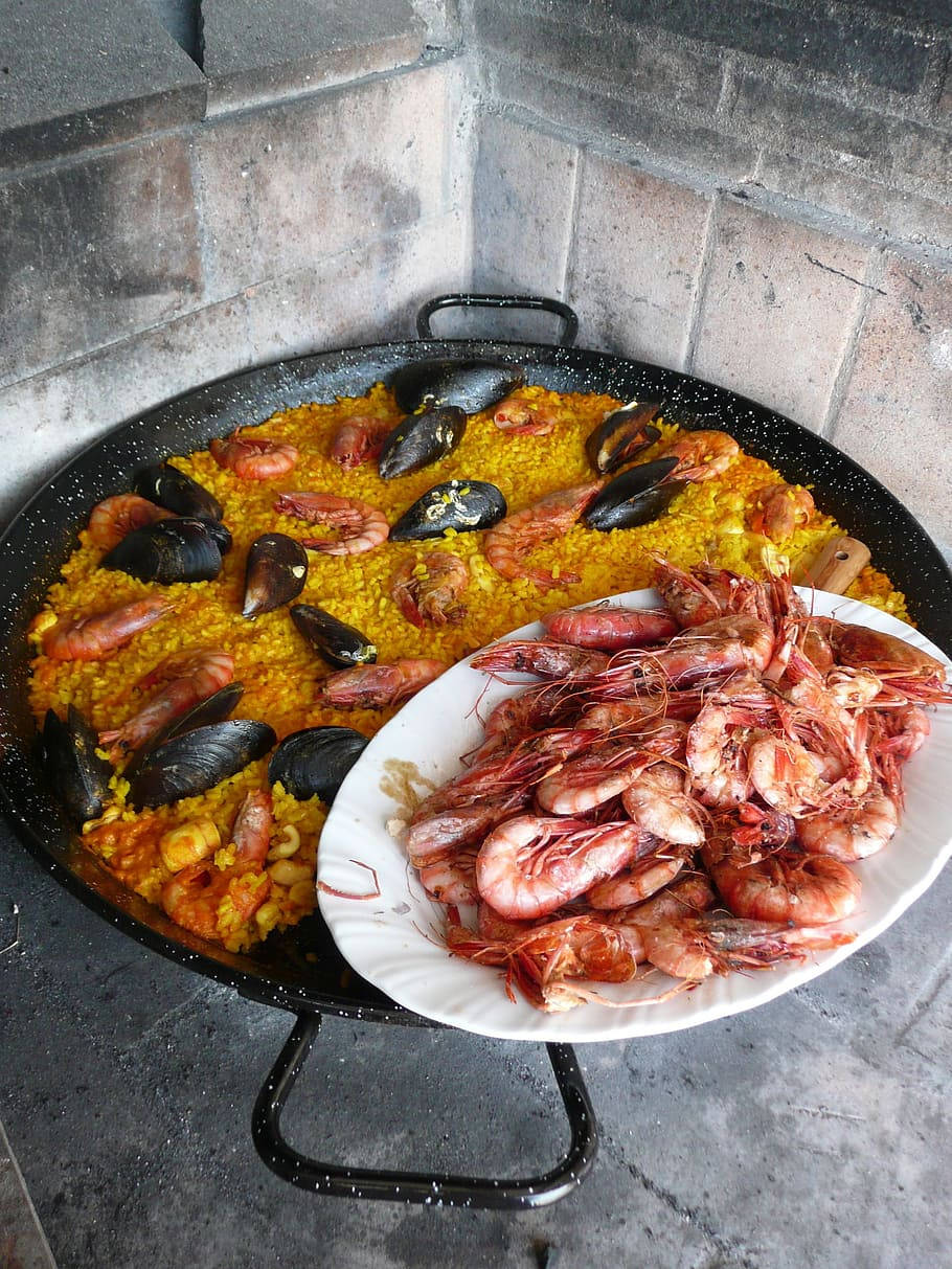 prawn 桌布 壁紙 — 一個大型西班牙海鮮飯鍋裝滿金黃色米飯和海鮮，旁邊放著一盤新鮮粉紅蝦，背景是灰色磚牆。