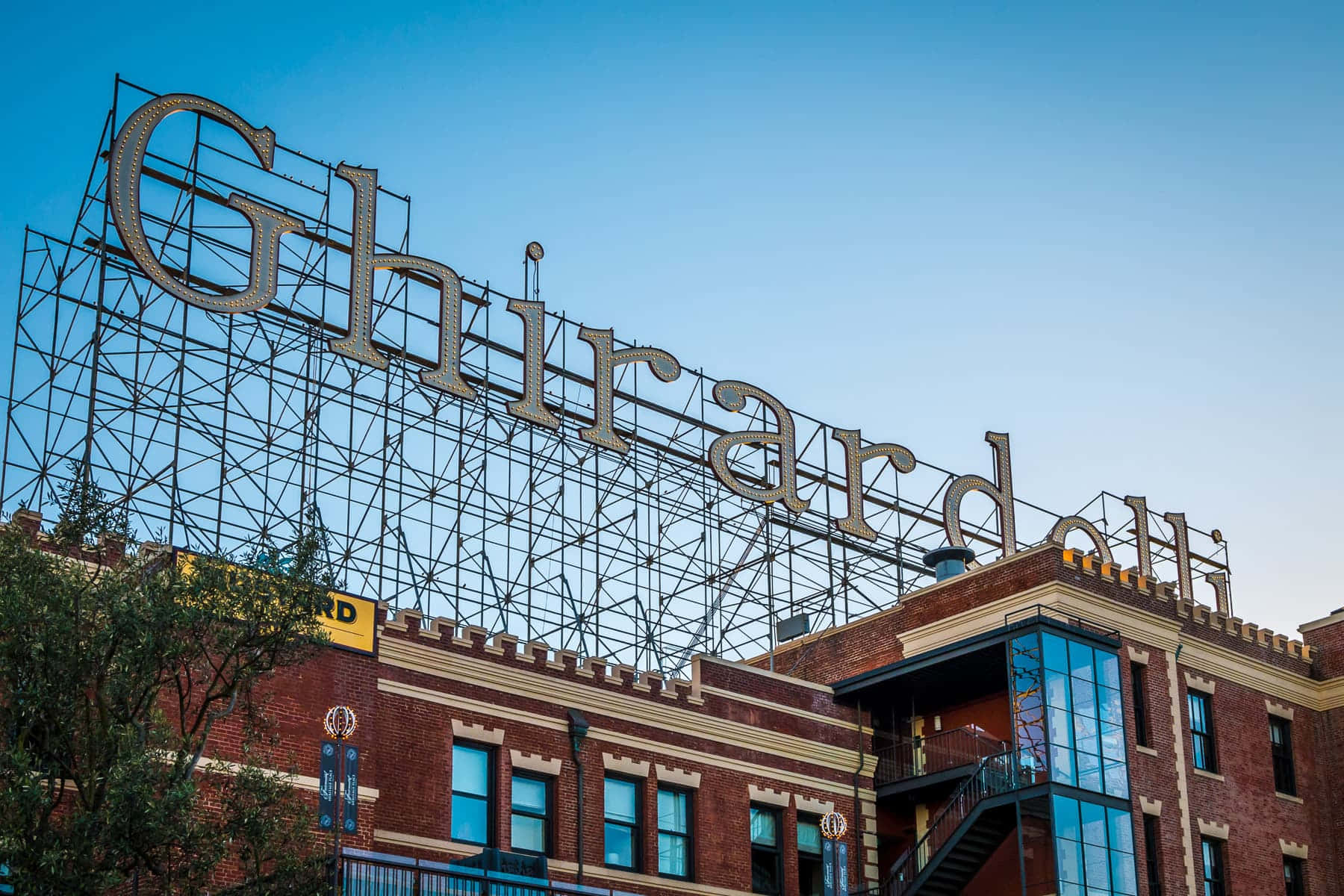 城市 桌布 壁紙 — 一個大型金屬 Ghirardelli 標誌聳立於紅磚建築之上，背景是清澈的藍天。