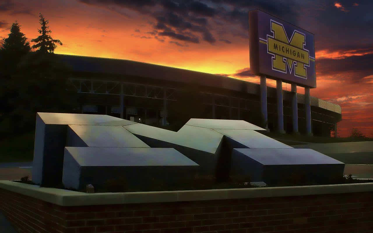 Michigan Football 桌布 壁紙 — 一個大型金屬M雕塑矗立在密西根體育場前，背景是火紅的夕陽天空。