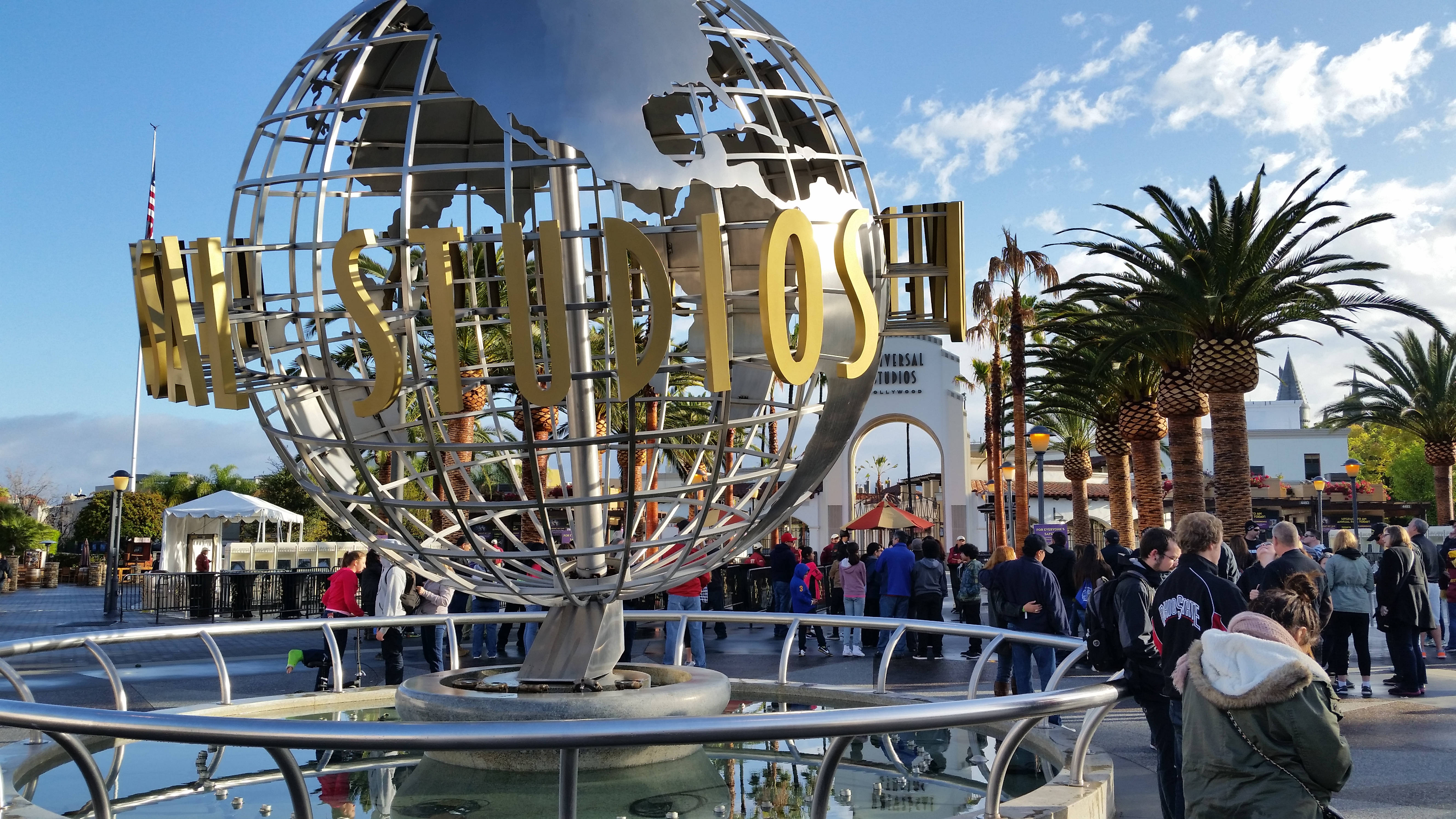 Universal 桌布 壁紙 — 一個大型金屬地球儀，上面有金色的「UNIVERSAL STUDIOS」字樣，矗立在主題公園入口處，藍天白雲，周圍有棕櫚樹和人群。