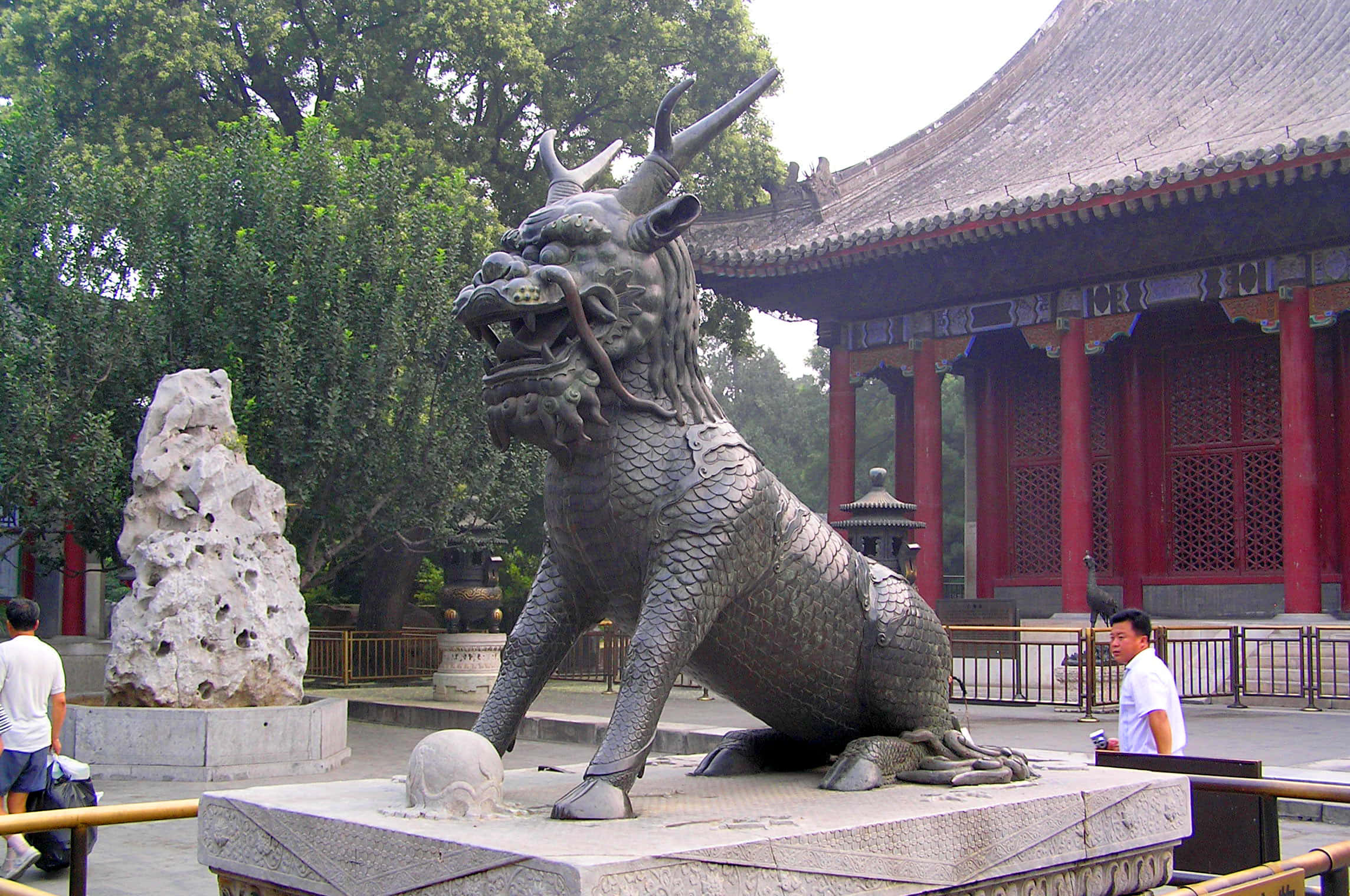 Statue Aesthetic 桌布 壁紙 — 一個大型金屬麒麟雕像守護著傳統中國寺廟庭院，庭院內有紅色柱子和茂密的綠色樹木。