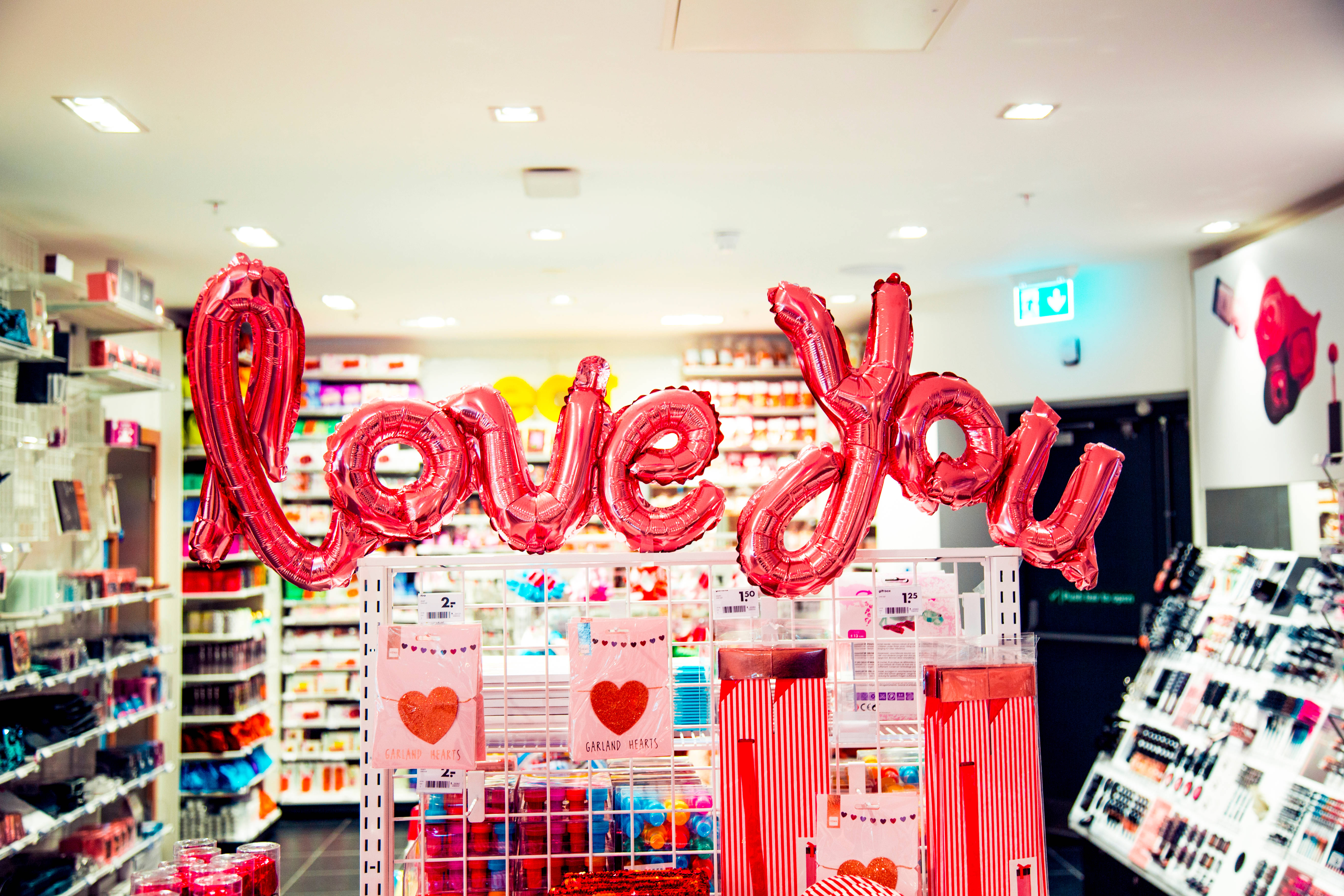 love you 桌布 壁紙 — 一個大型閃亮紅色 'Love You' 氣球漂浮在光線明亮的商店通道中的情人節展示上方。