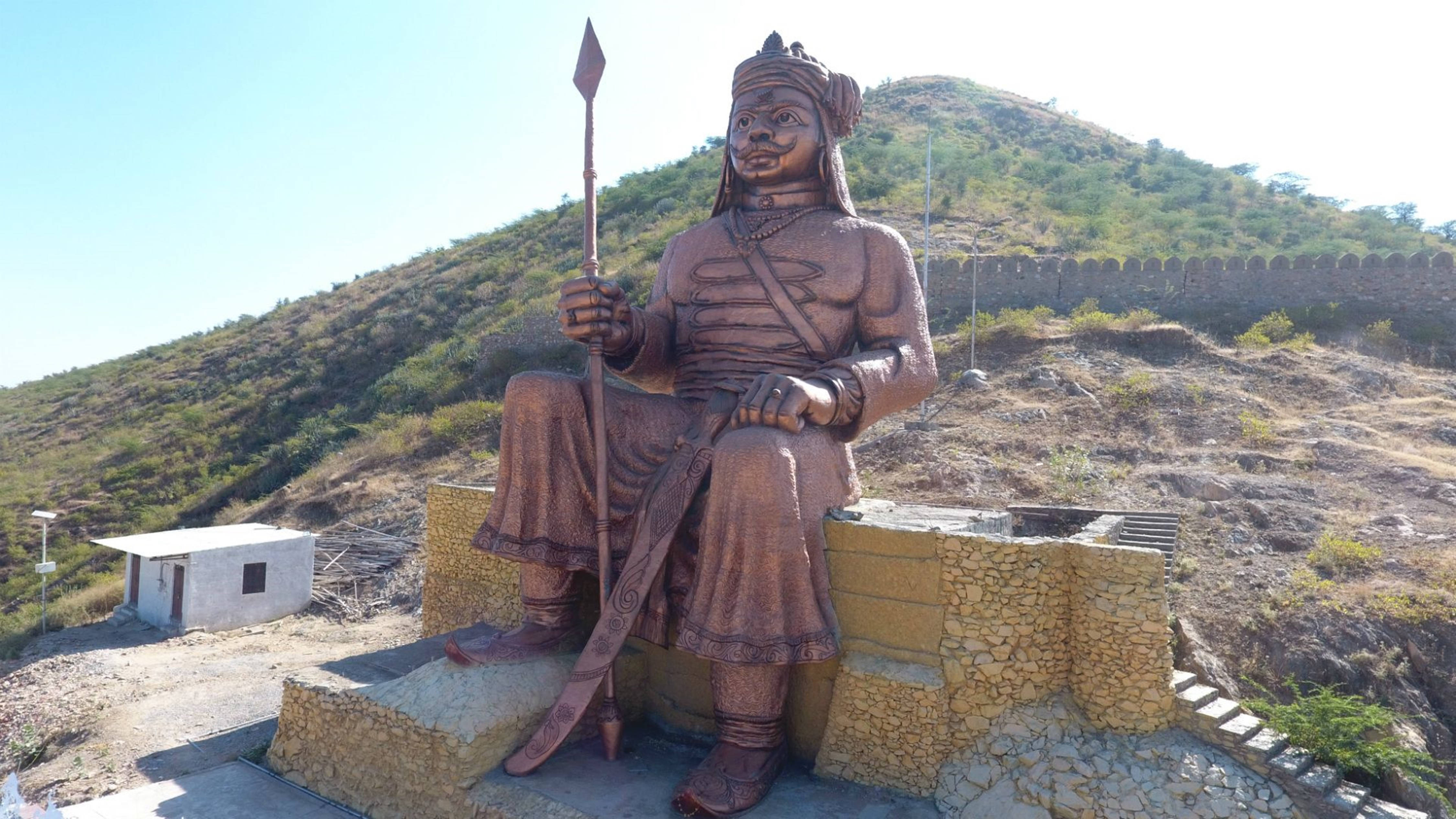 hill view 桌布 壁紙 — 一個大型青銅雕像，描繪一位坐著的戰士手持長矛和劍，立於石基座上，背景是陽光照耀的山坡和遠處的牆壁。
